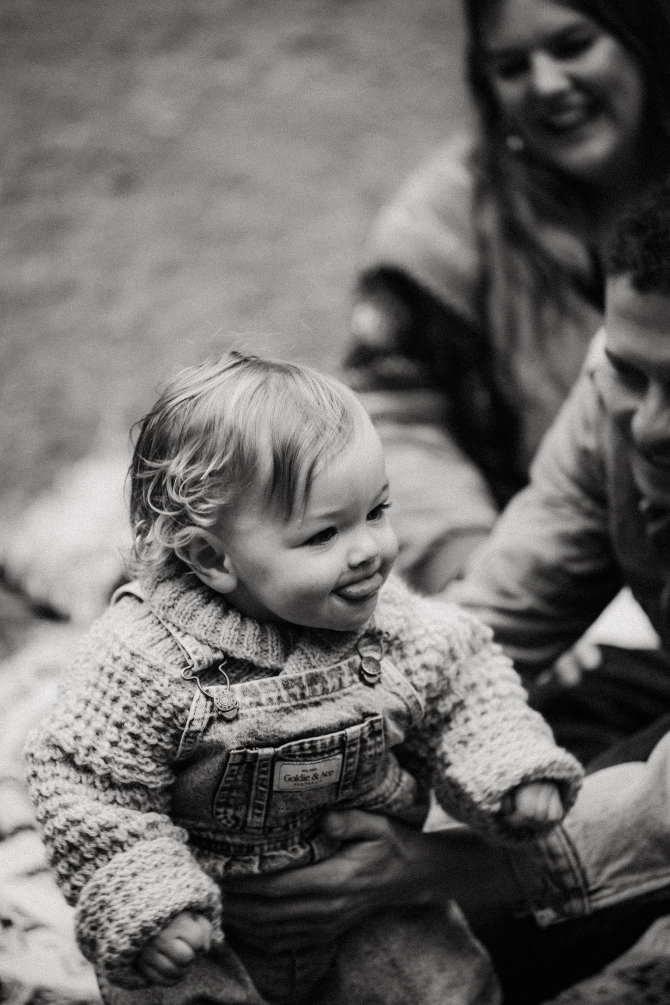 Black and white photo of a smiling toddler in knit sweater and overalls, held by parents sitting outdoors.
