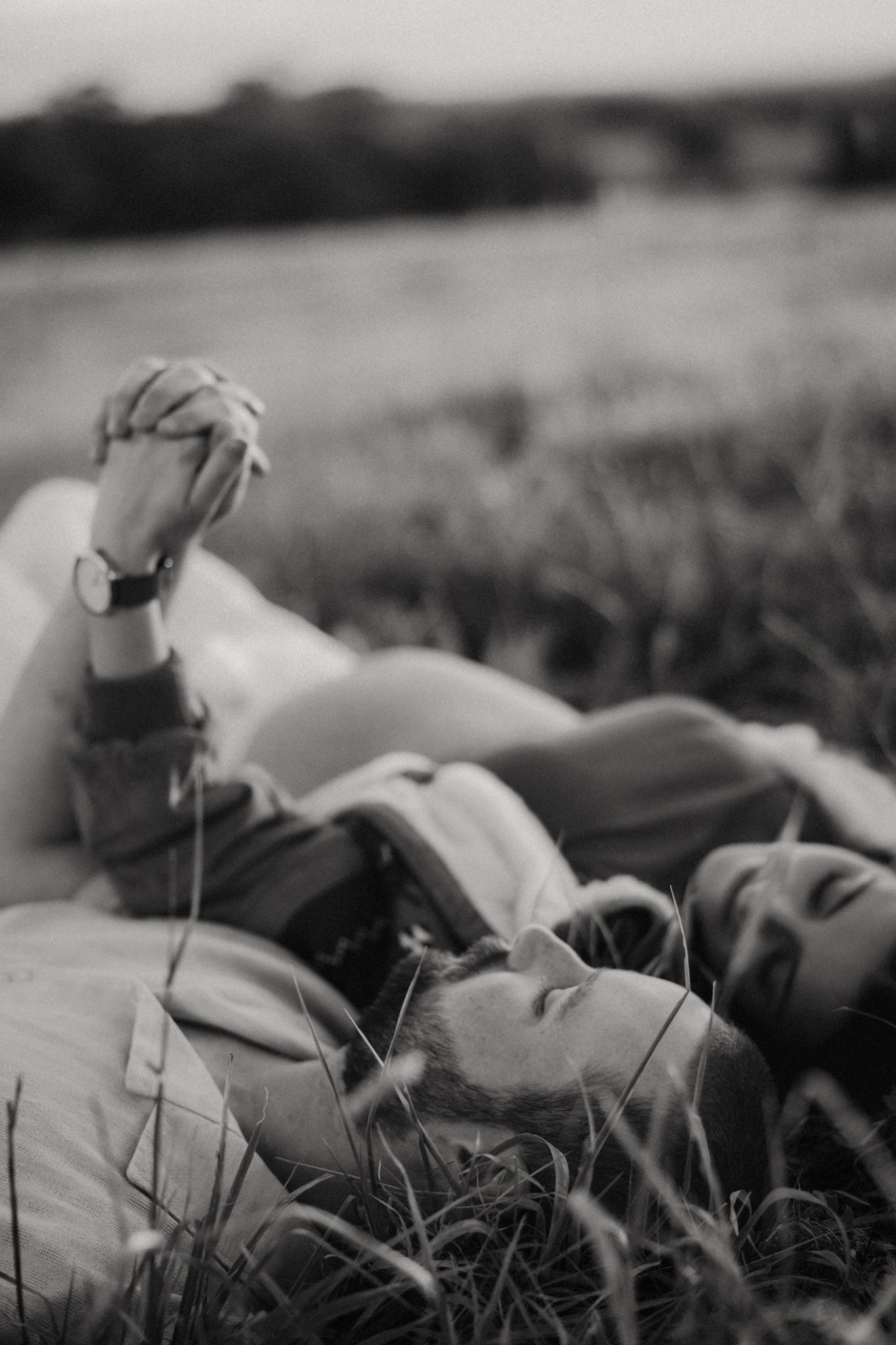 Black and white photo of a couple lying in grass, holding hands, with a blurred field in the background.