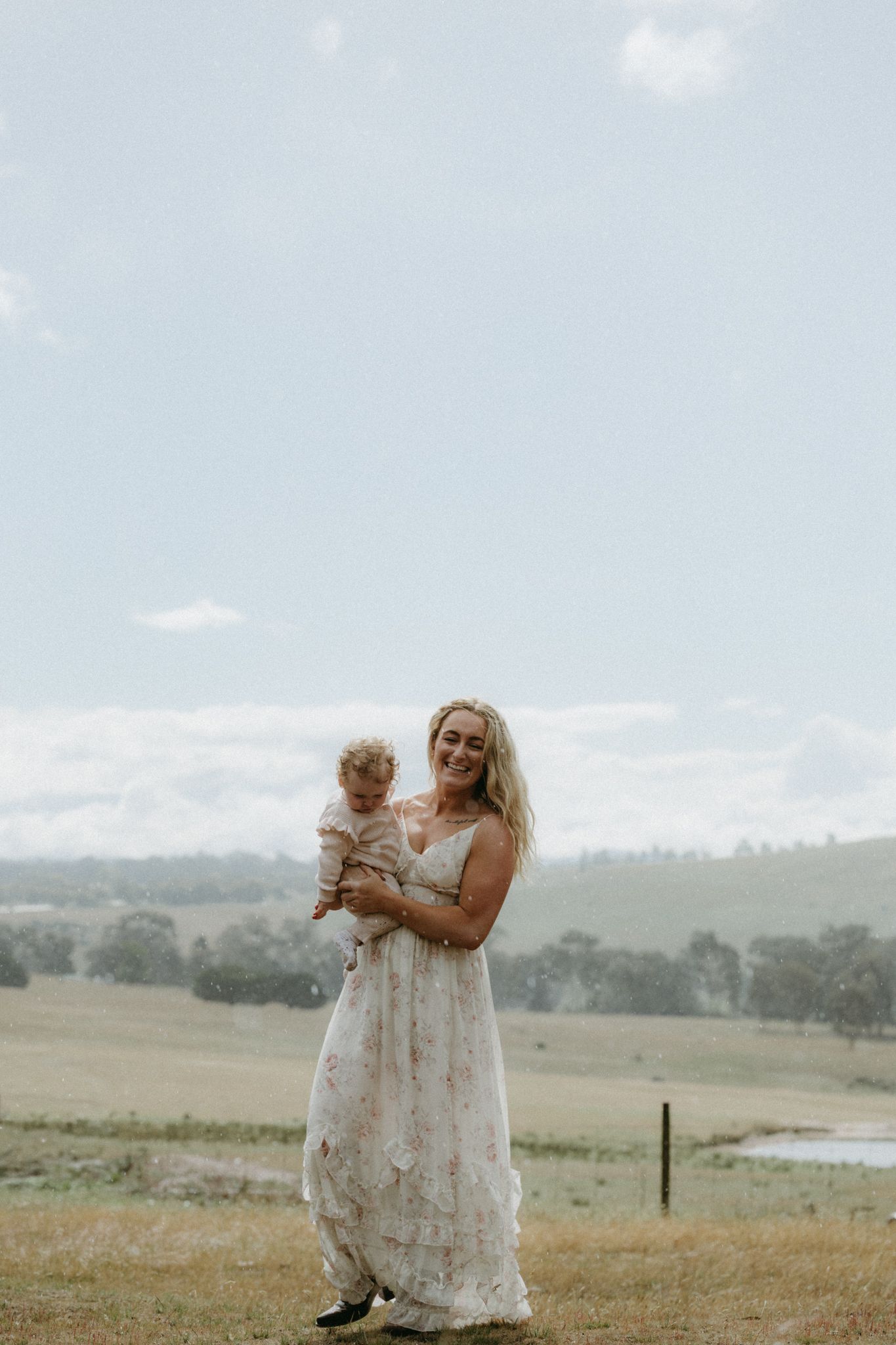 Smiling blonde woman in floral maxi dress holding a baby in a scenic green countryside setting.