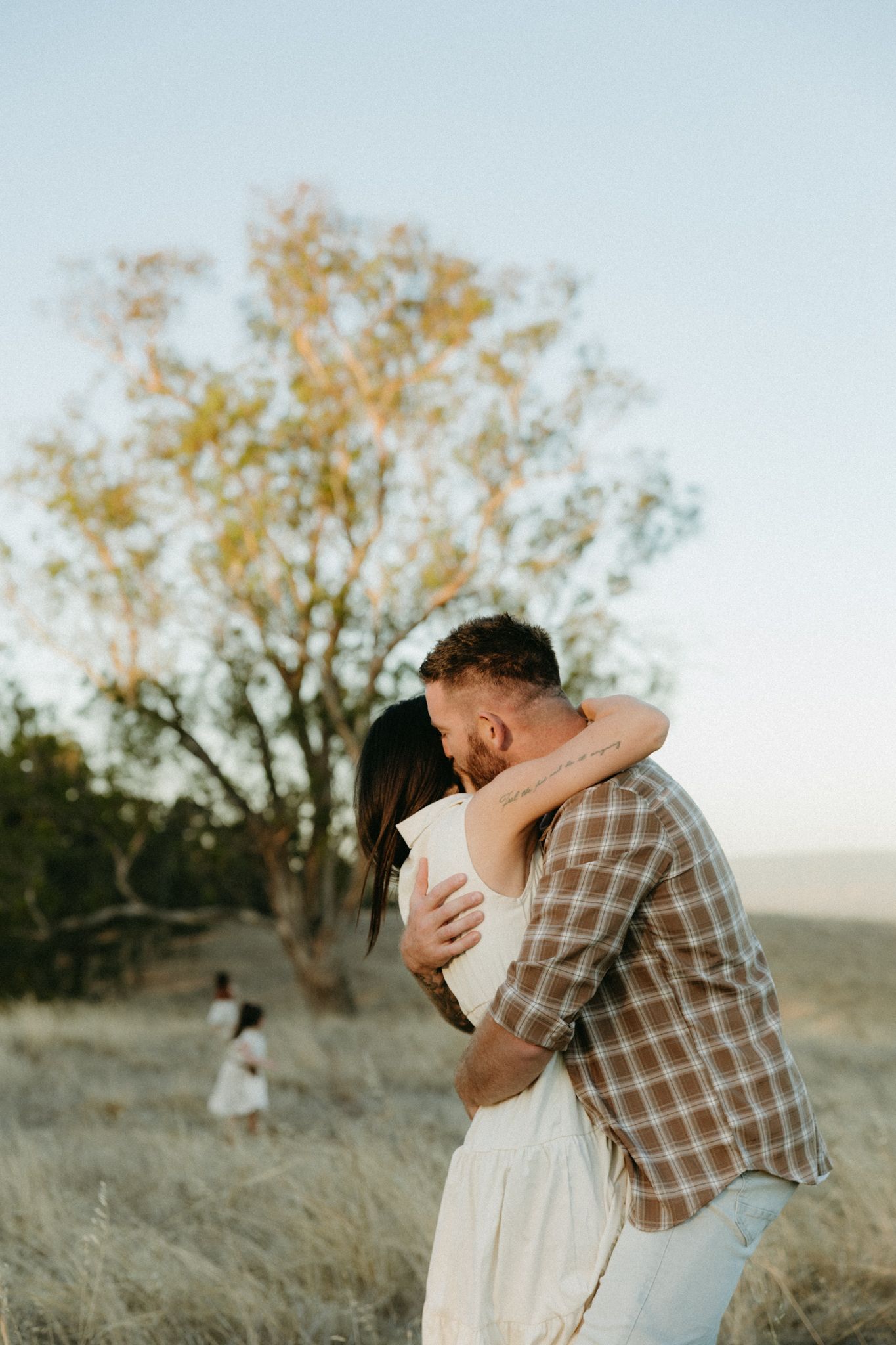 Couple embracing romantically in a golden field at sunset with a tree and child in the background.