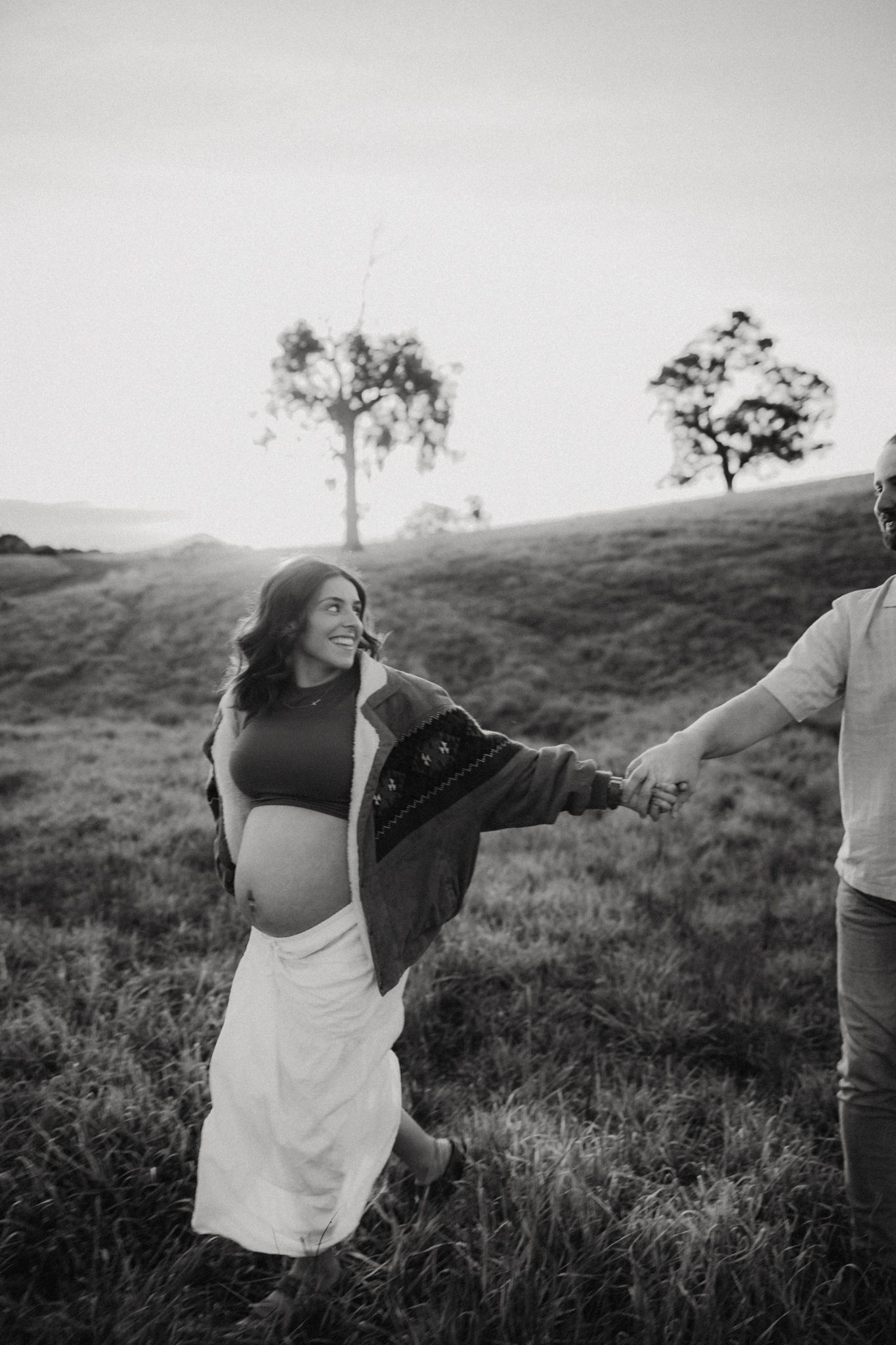 Black and white maternity photo of smiling pregnant woman holding hands with partner in grassy field at sunset.