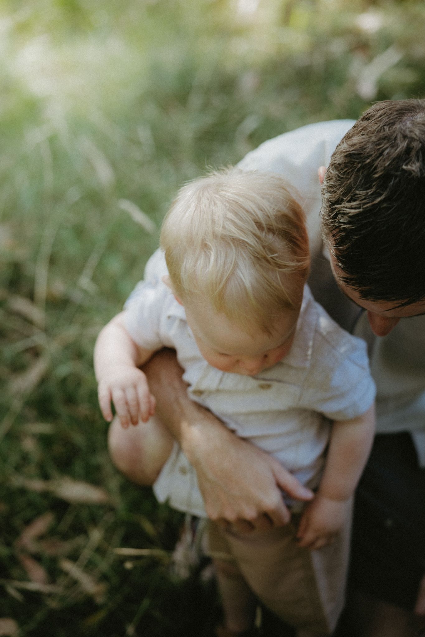 A father crouches down embracing his blonde toddler outdoors in a lush green field.