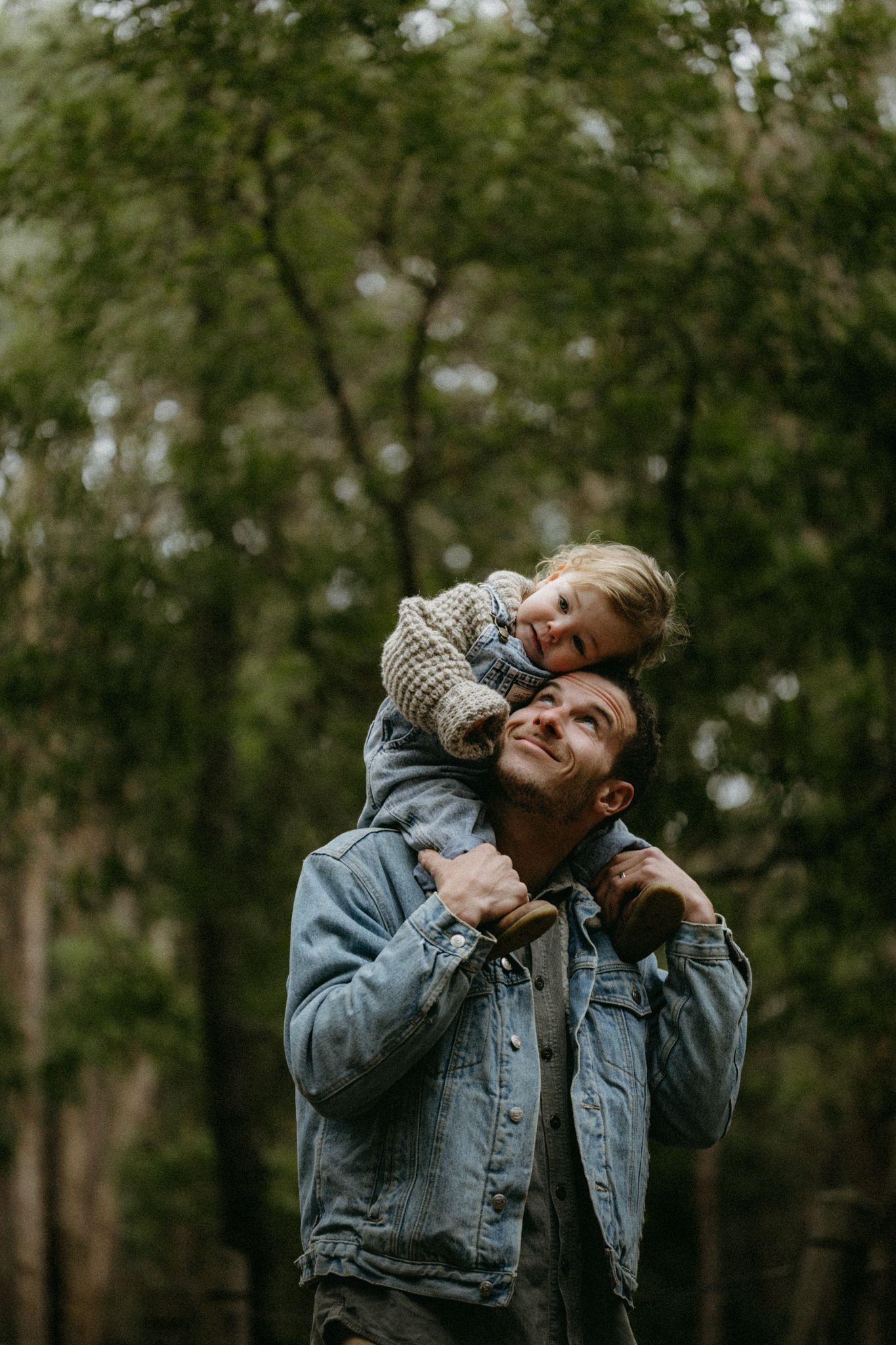 A smiling man in a denim jacket carries a happy toddler on his shoulders in a lush green forest setting.