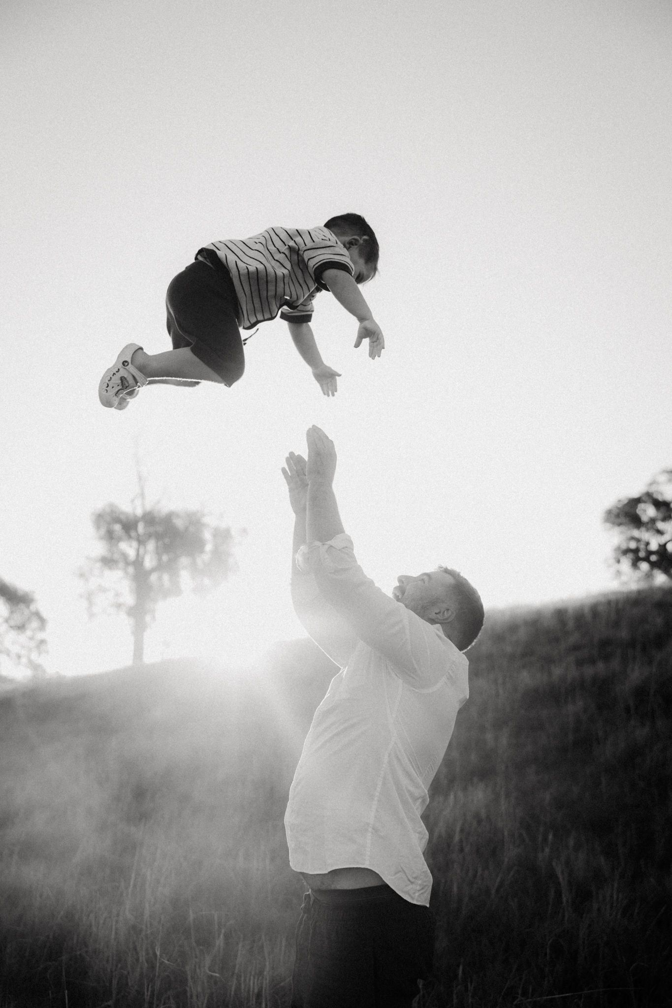Black and white photo of a father tossing his young son high in the air outdoors at sunset.