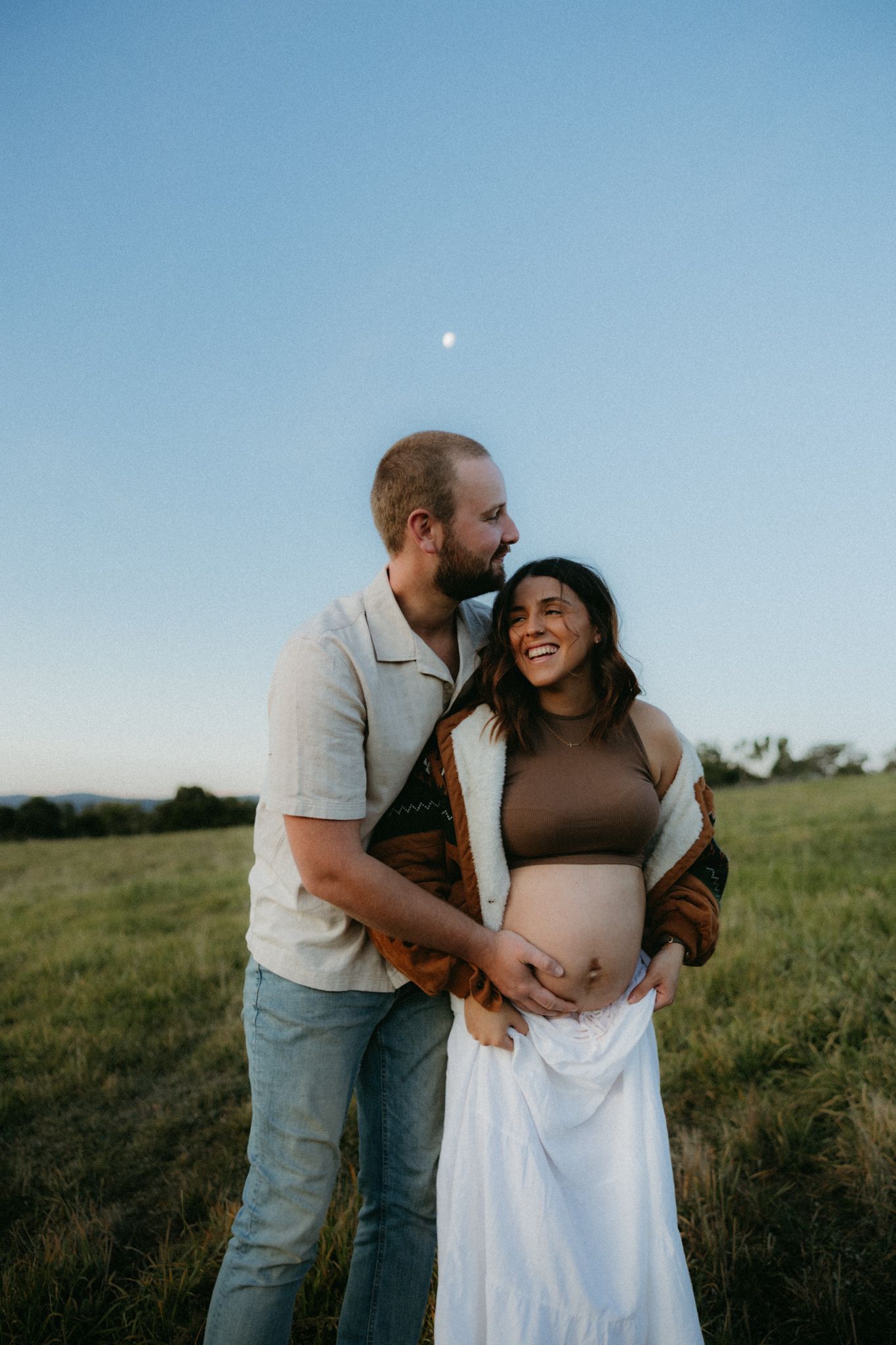 Happy pregnant couple embracing in a green field at dusk, woman laughing joyfully while partner holds her baby bump.