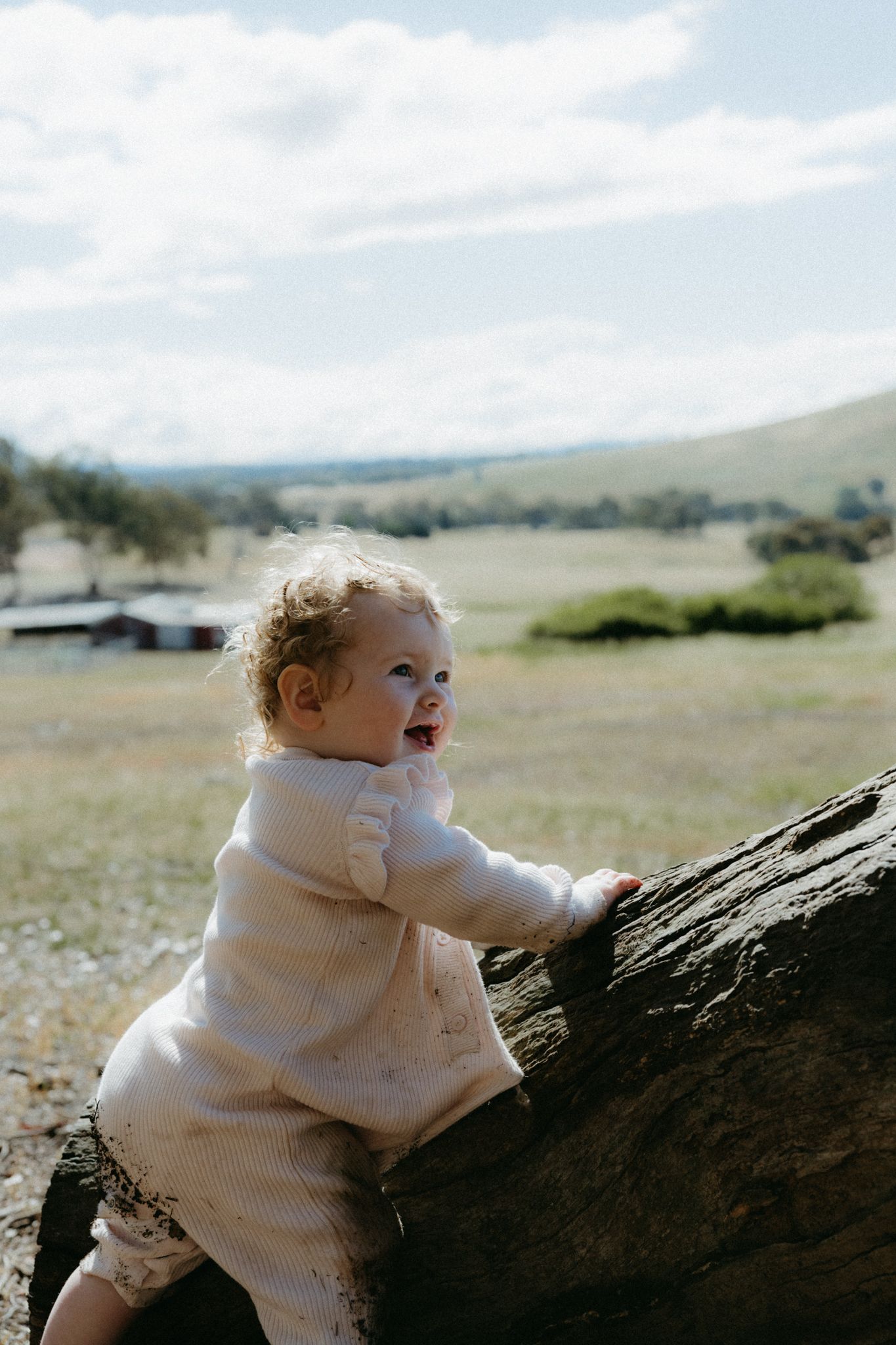 Toddler in white outfit climbing a large rock in a scenic green countryside landscape under a cloudy sky.