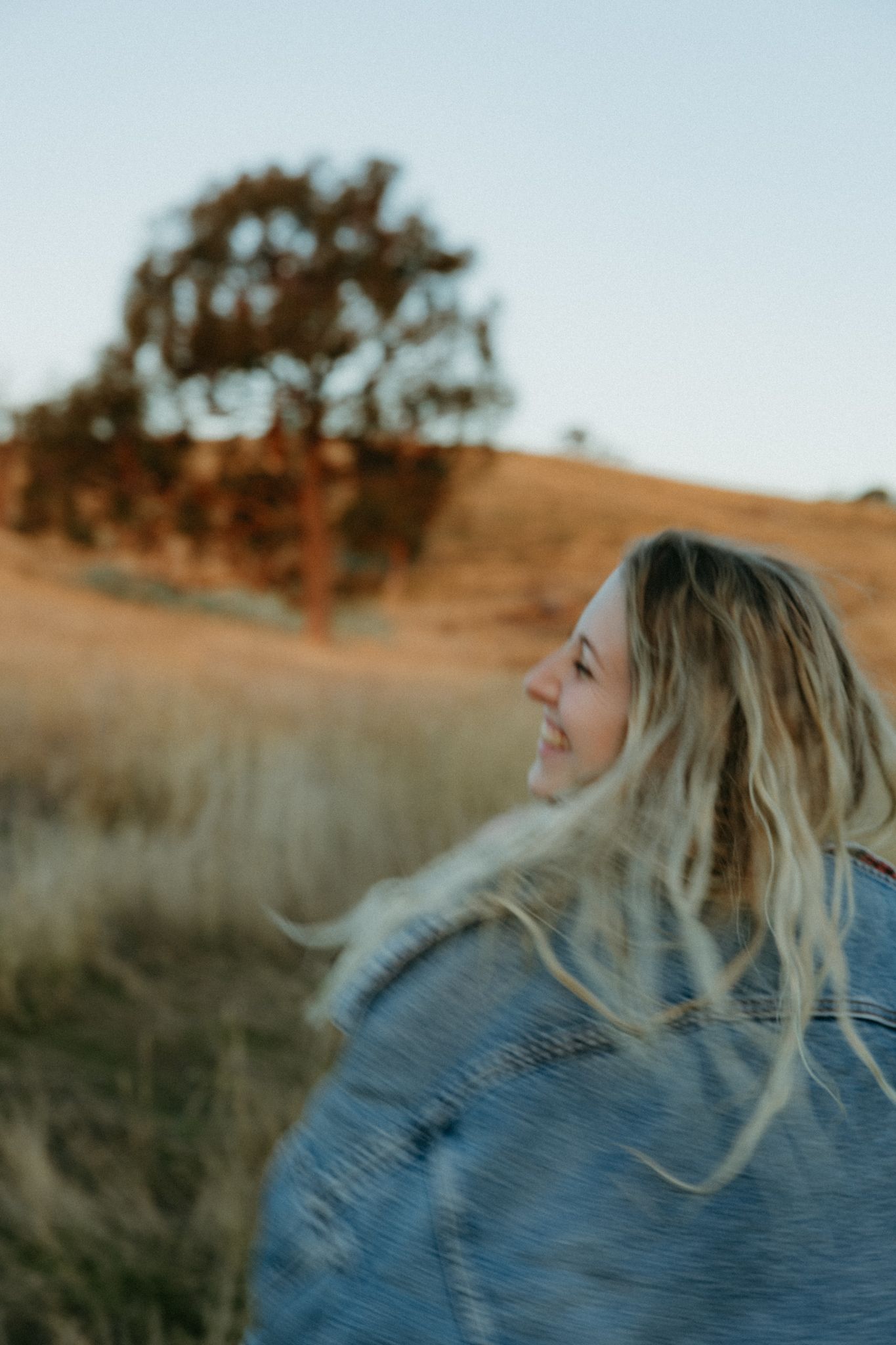 Smiling blonde woman in denim jacket with hair blowing in wind, golden field and tree in background at sunset.