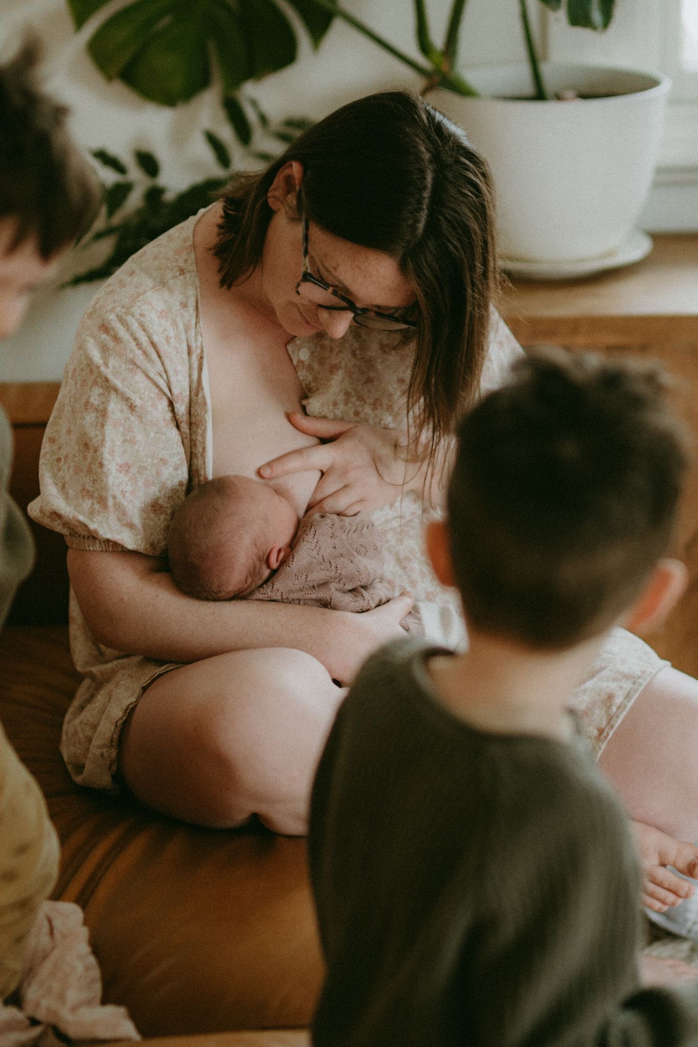 A mother breastfeeding her newborn baby while sitting on the floor, surrounded by older children and indoor plants.