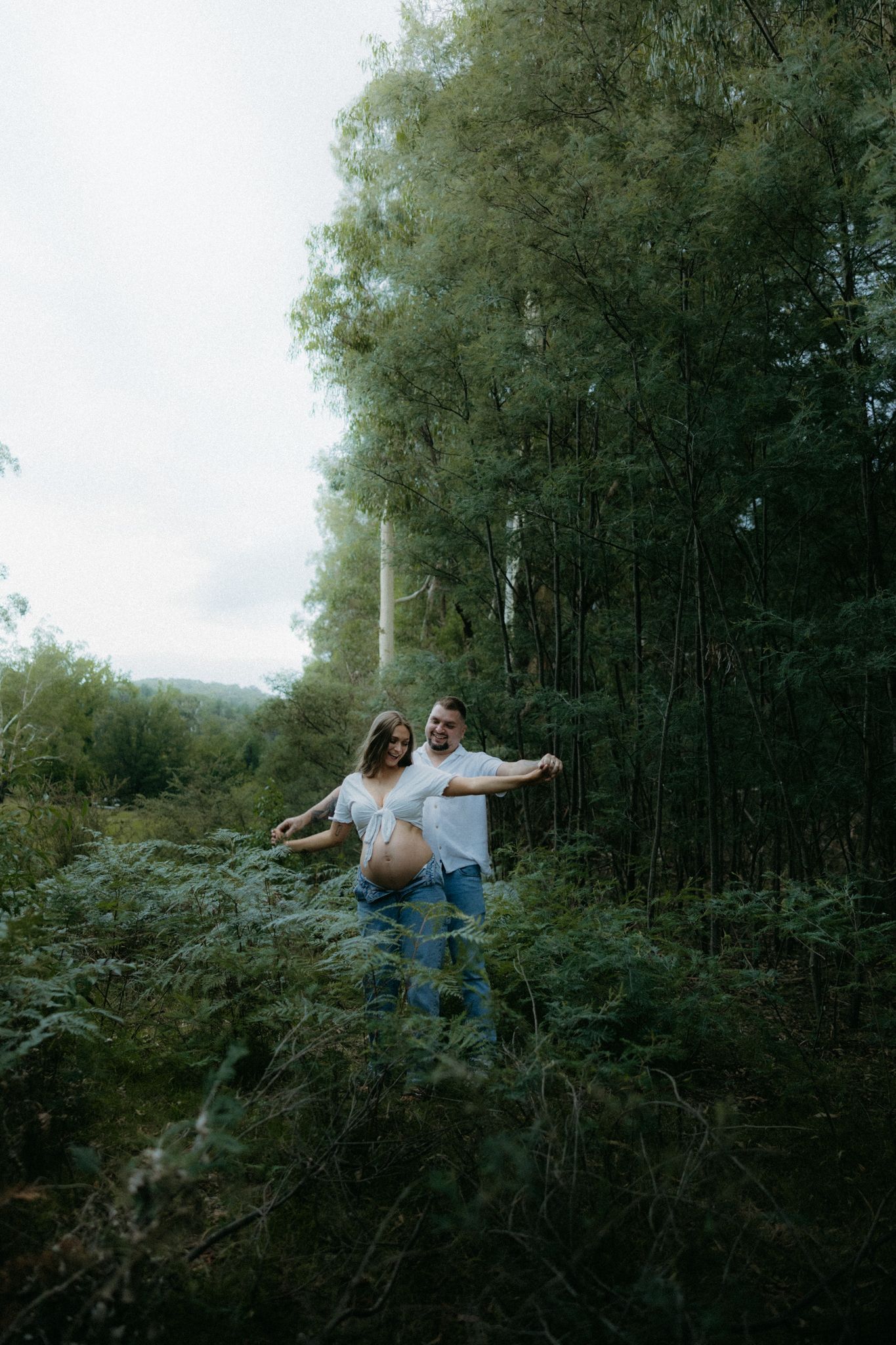 Pregnant couple posing together in lush green forest, woman showing baby bump, surrounded by ferns and tall trees.
