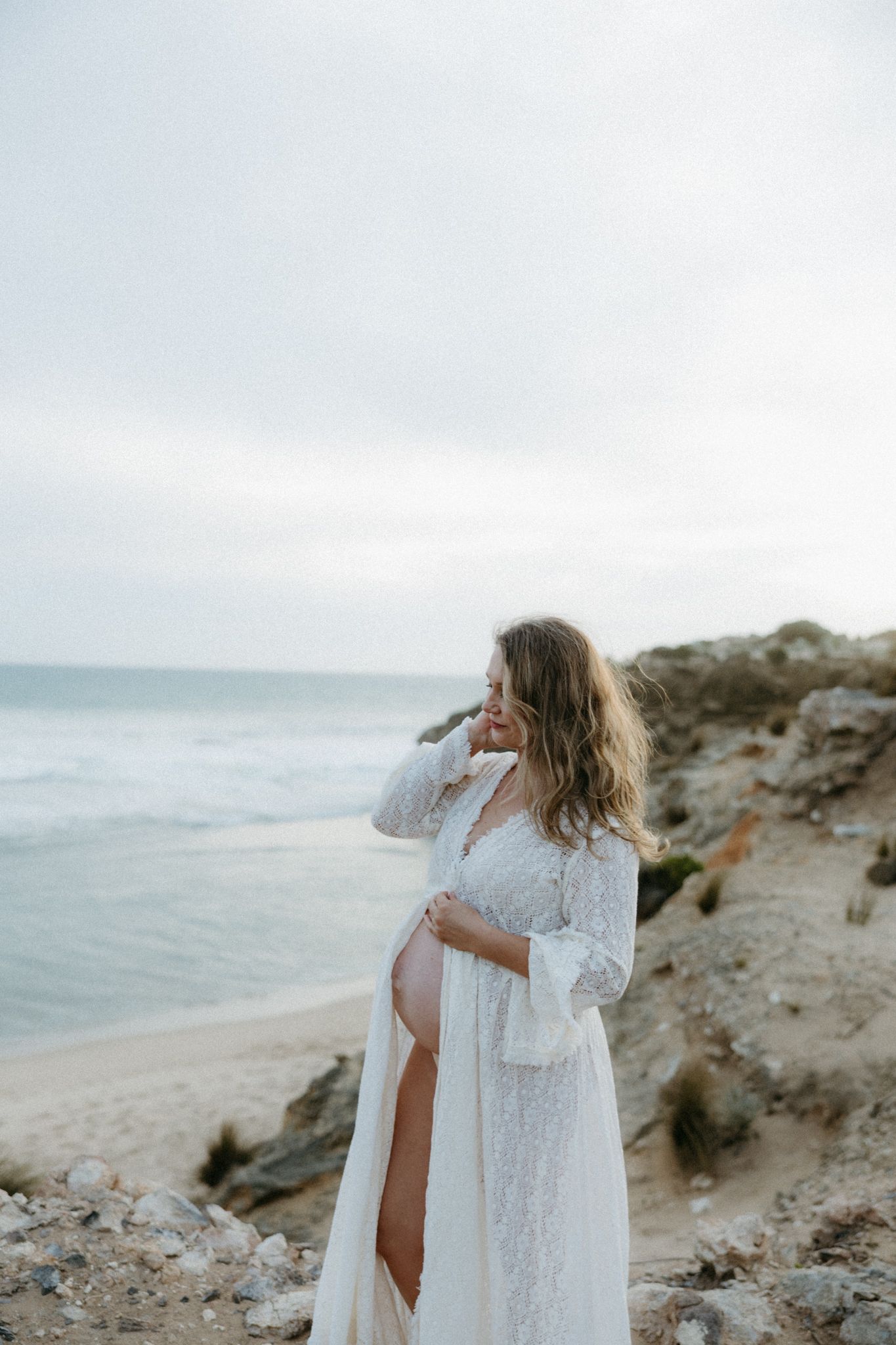 Pregnant woman in white lace robe cradling her baby bump on a rocky beach with ocean waves behind her.