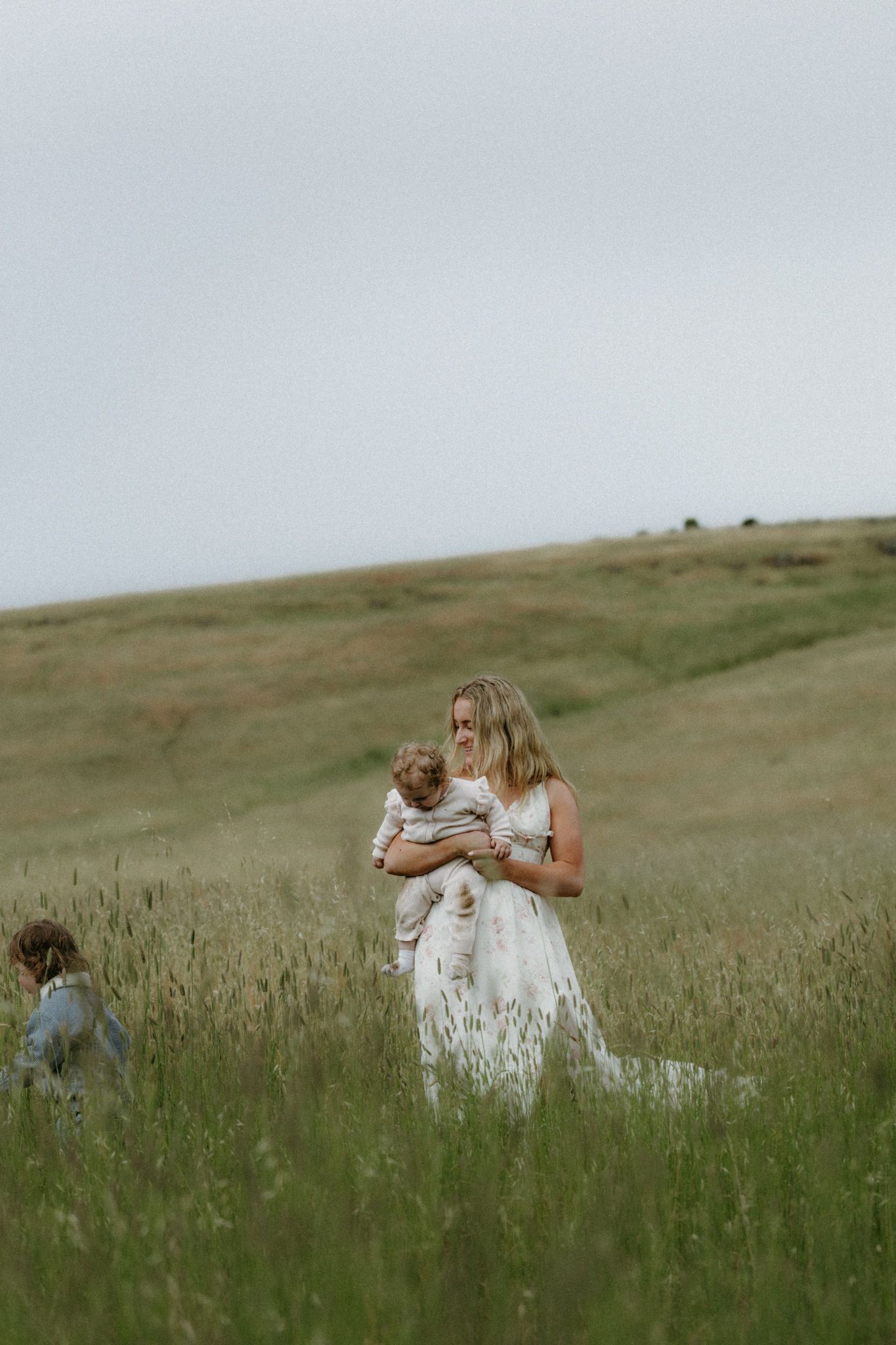 Woman in white dress holding a baby in a green meadow field with a child nearby under overcast skies.
