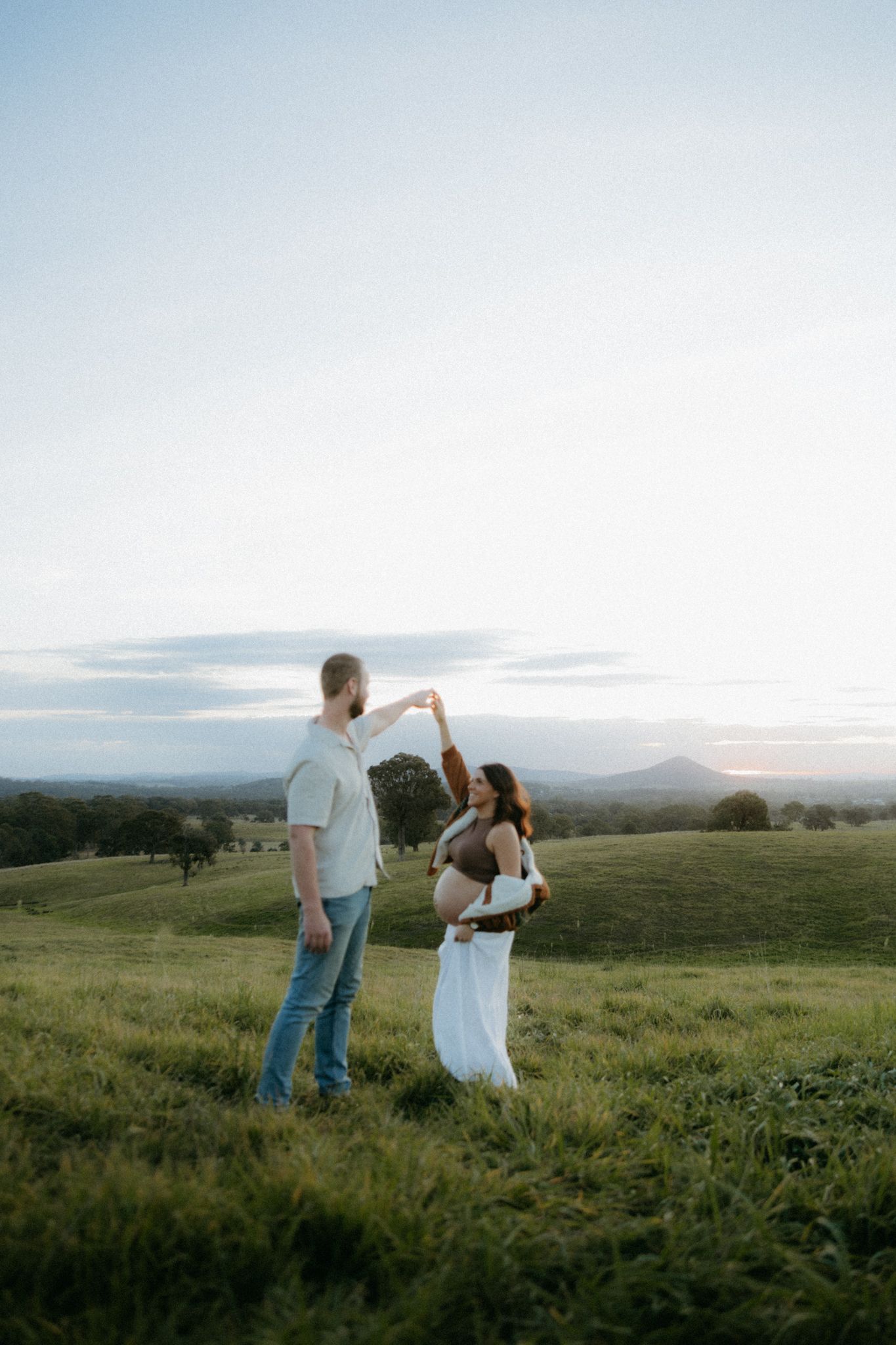 Pregnant couple dancing in green rolling hills at sunset, man spinning woman in white maternity dress outdoors.