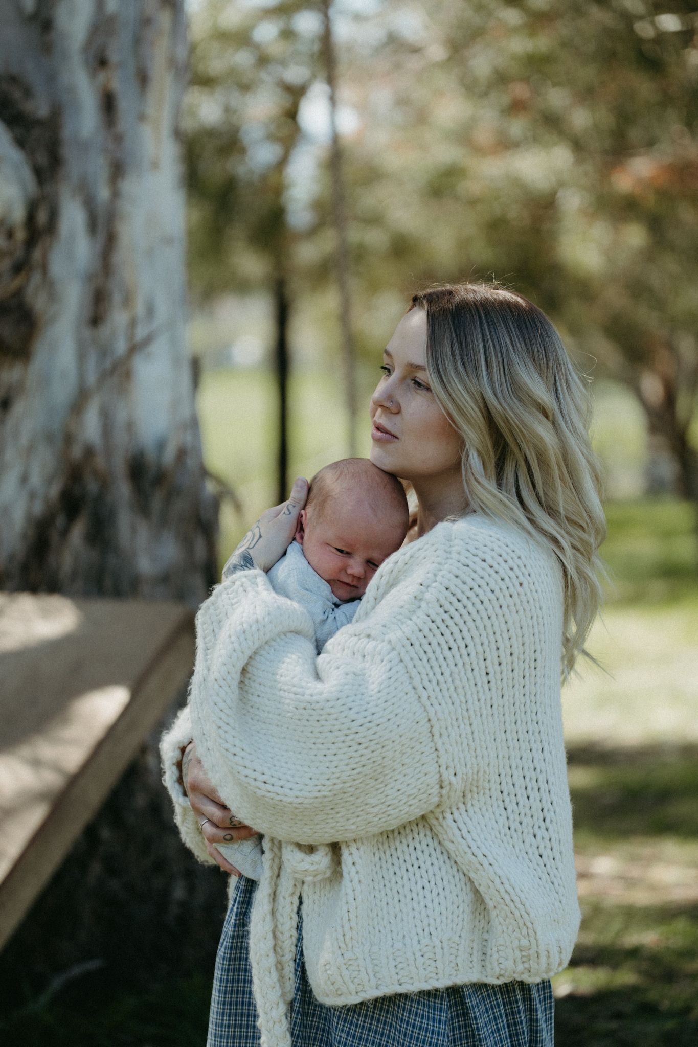 A young blonde mother in a cream knit sweater tenderly holds her sleeping newborn baby outdoors in a park.