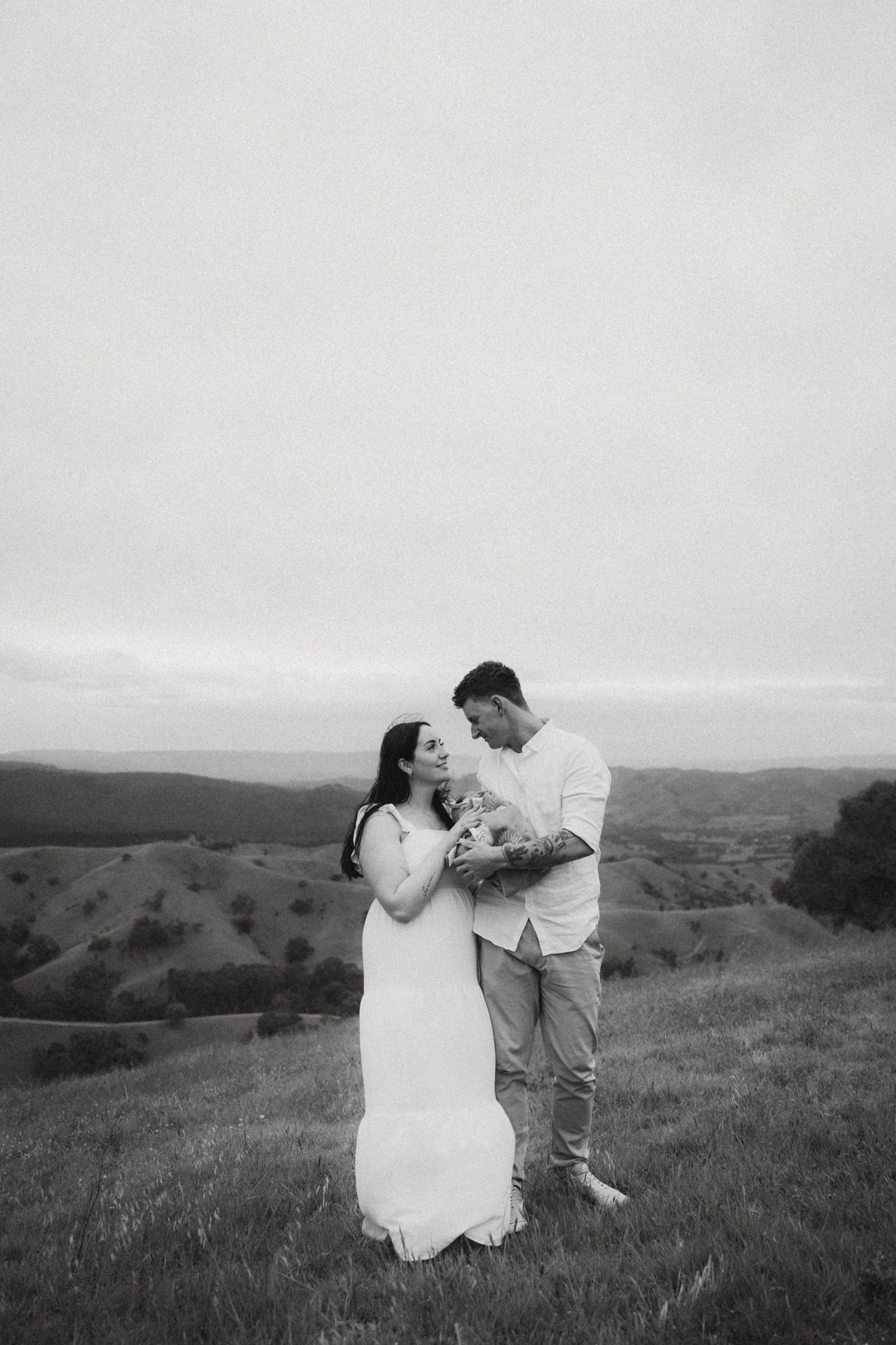 Black and white photo of a couple embracing on a hillside meadow, woman in white dress holding flowers, rolling hills behind them.