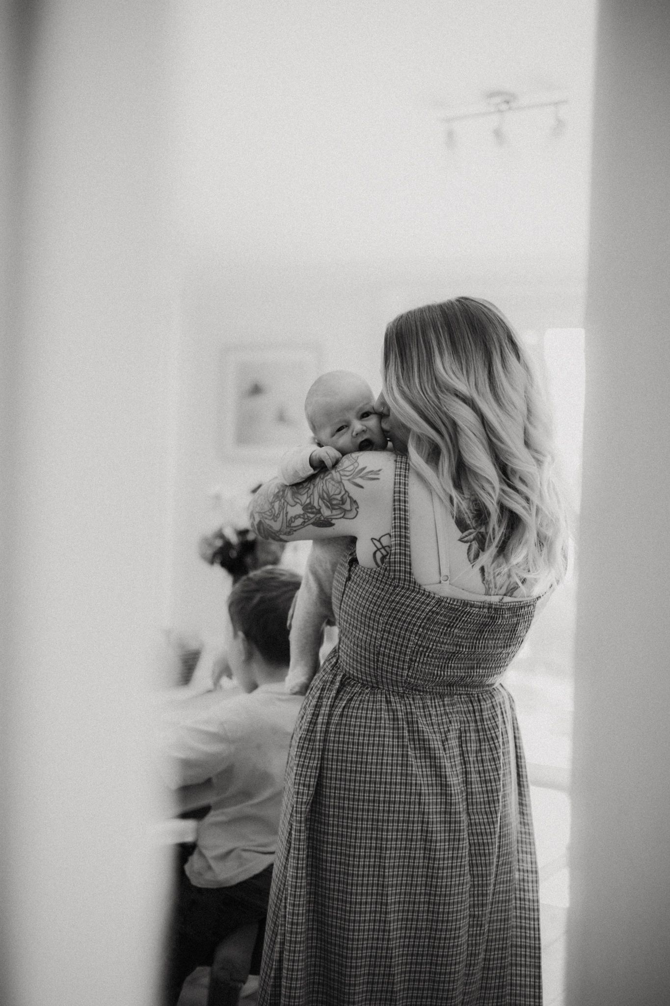 Black and white photo of a tattooed blonde mother holding her newborn baby near a bright window indoors.