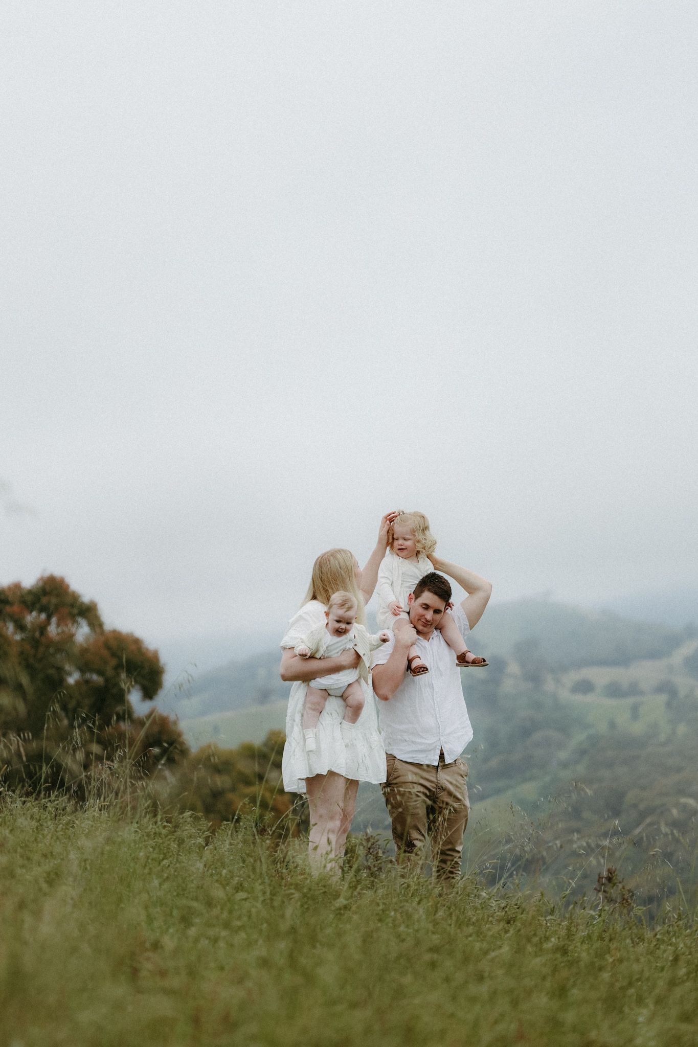 Family of four enjoying a misty green hillside, parents holding two young children in white outfits with rolling hills behind.