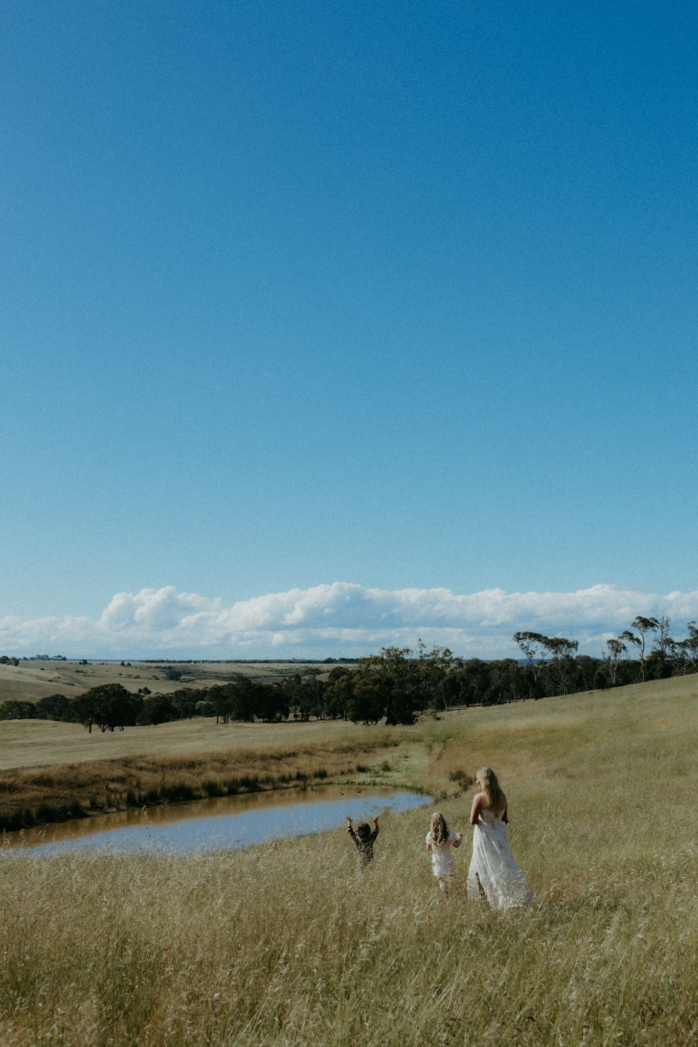 Woman and children walking through golden fields near a pond under a bright blue sky on a sunny day.