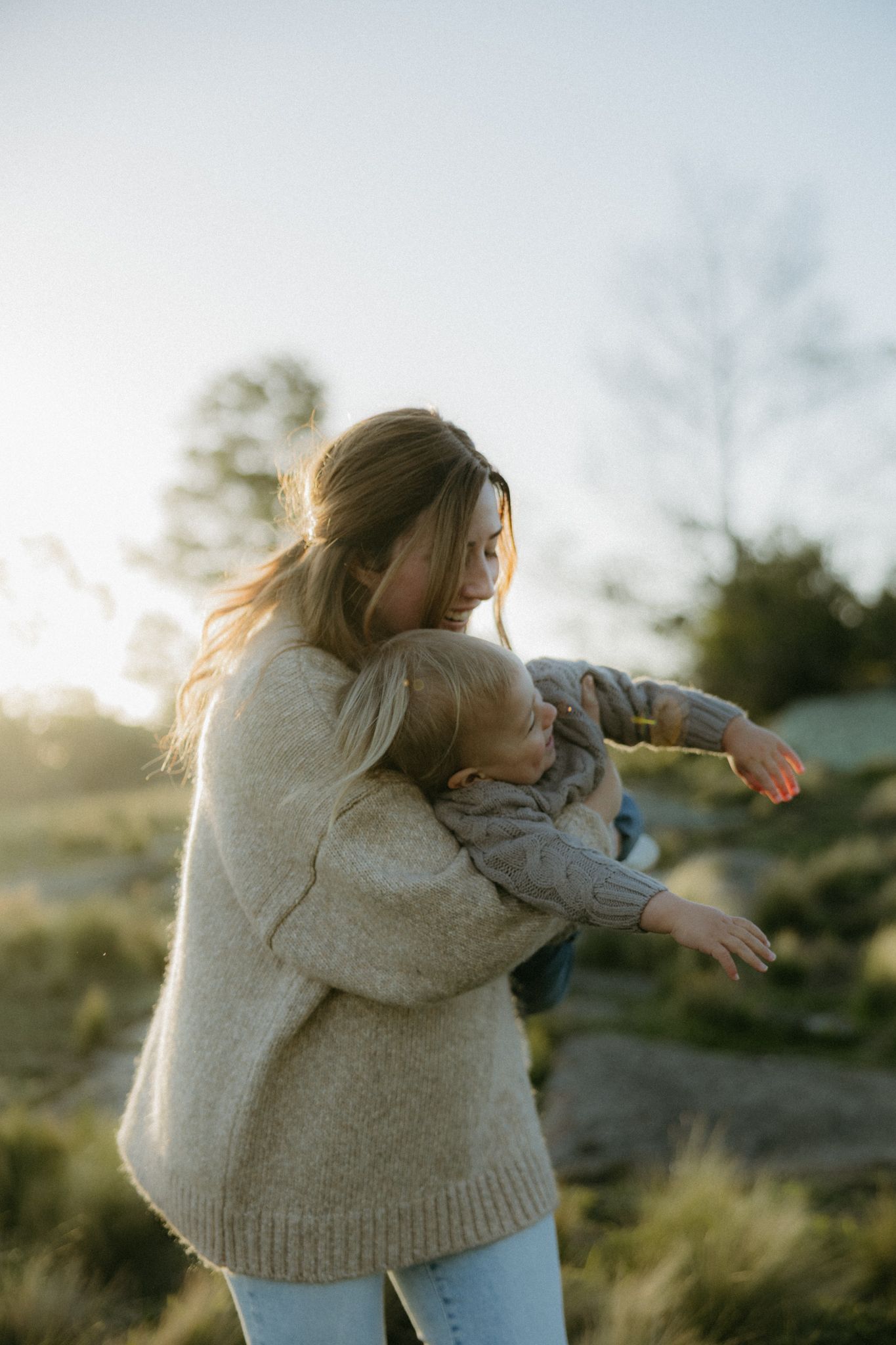 A mother tenderly holds her young child outdoors at golden hour, both wearing cozy sweaters in a natural setting.