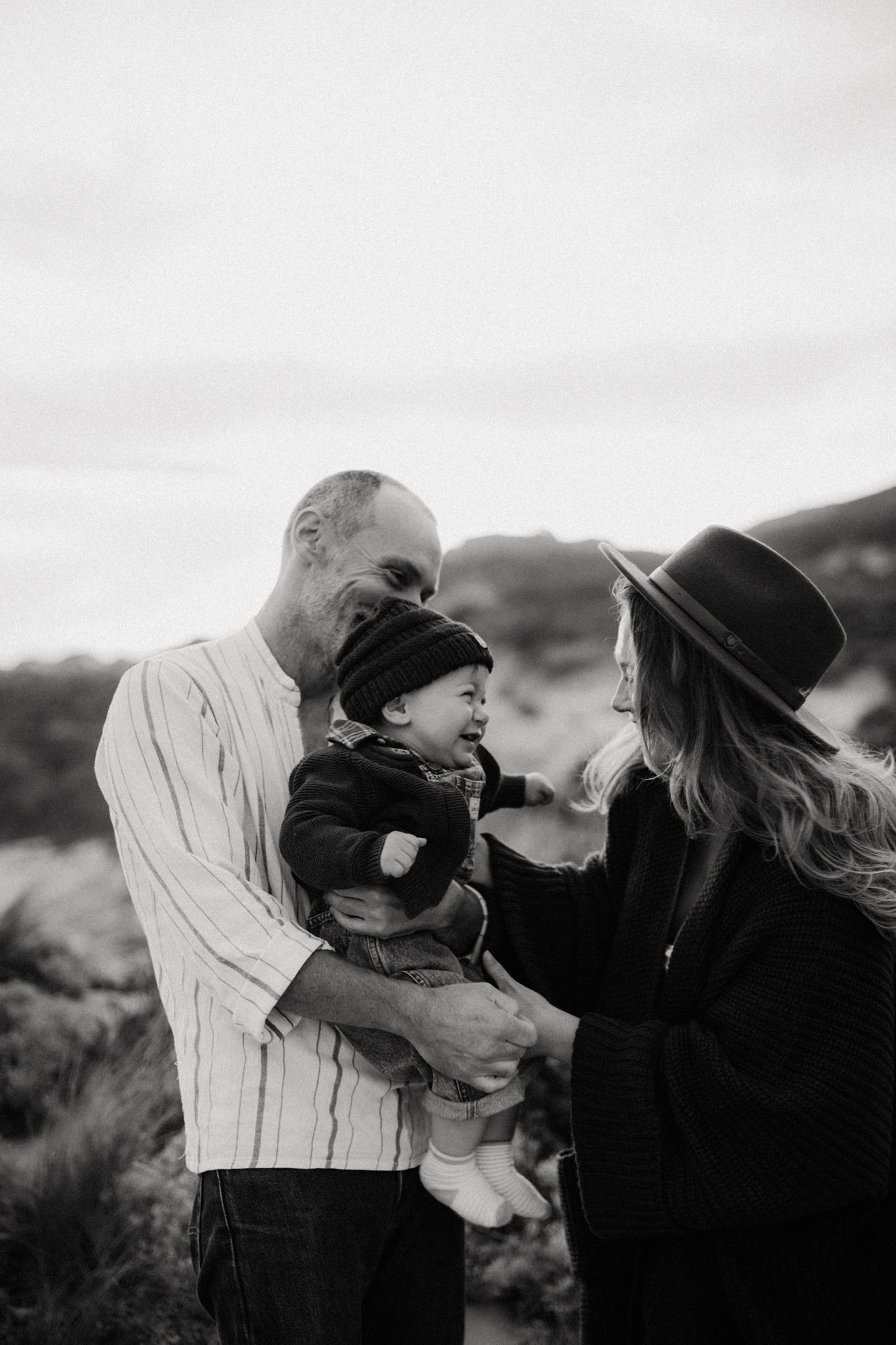 Black and white photo of a bald man and woman in hat lovingly holding a baby outdoors in a natural landscape.
