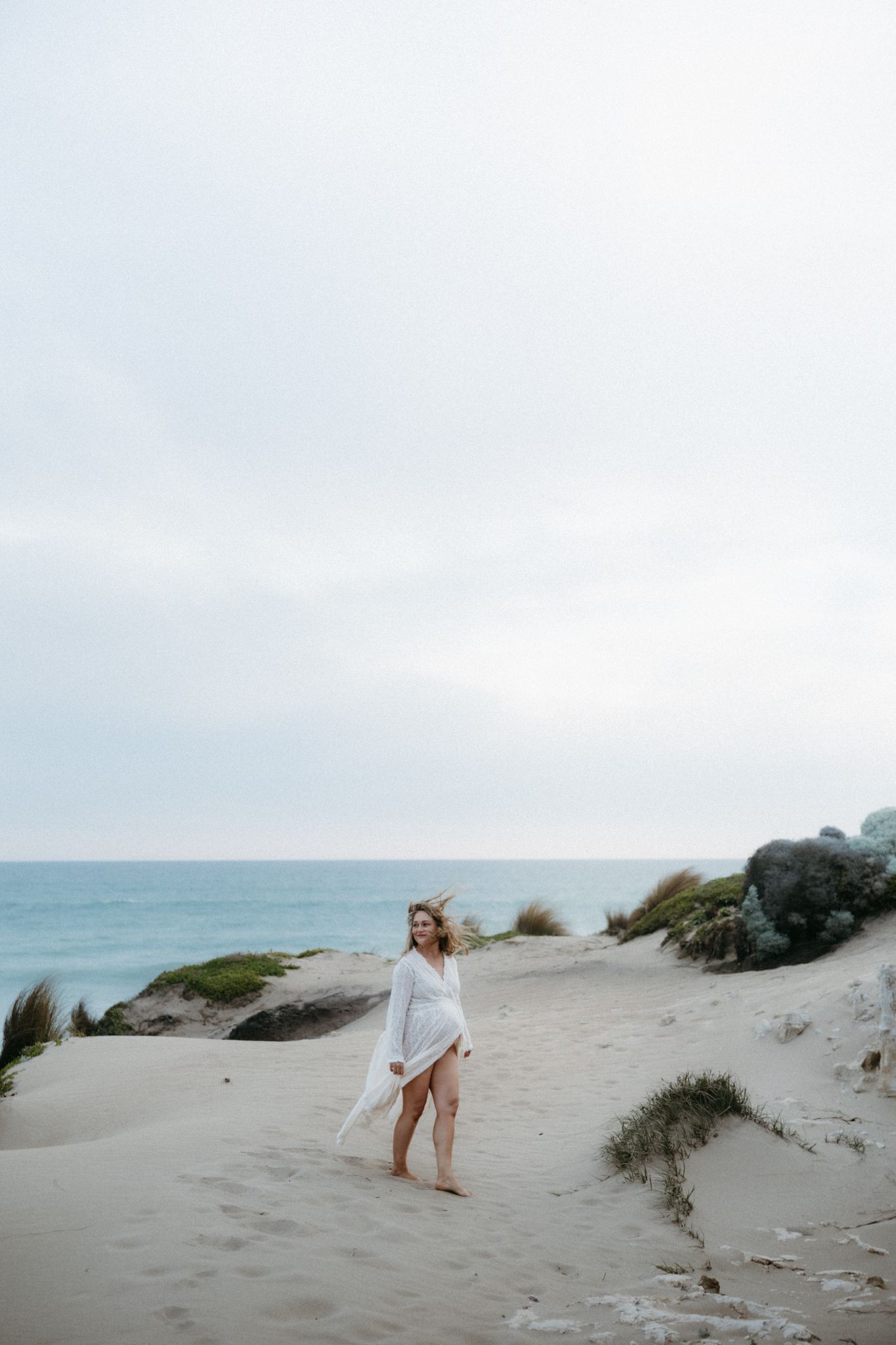 A woman in a flowing white dress walks barefoot on sandy beach dunes with ocean and rocks in background.
