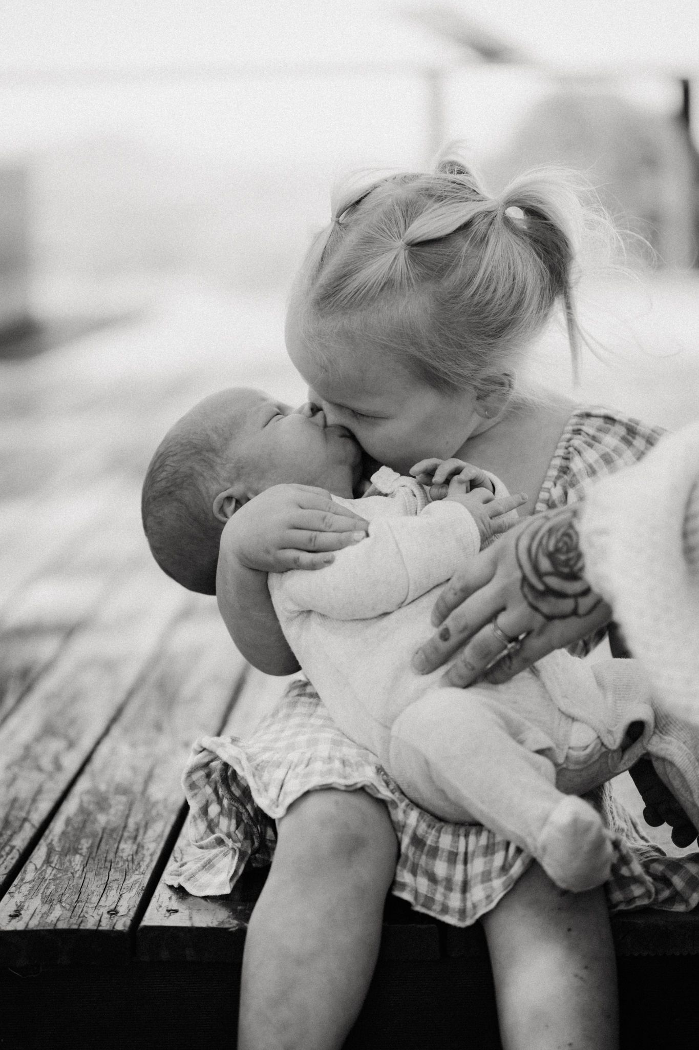 Black and white photo of a young girl kissing a newborn baby while sitting on a wooden bench.