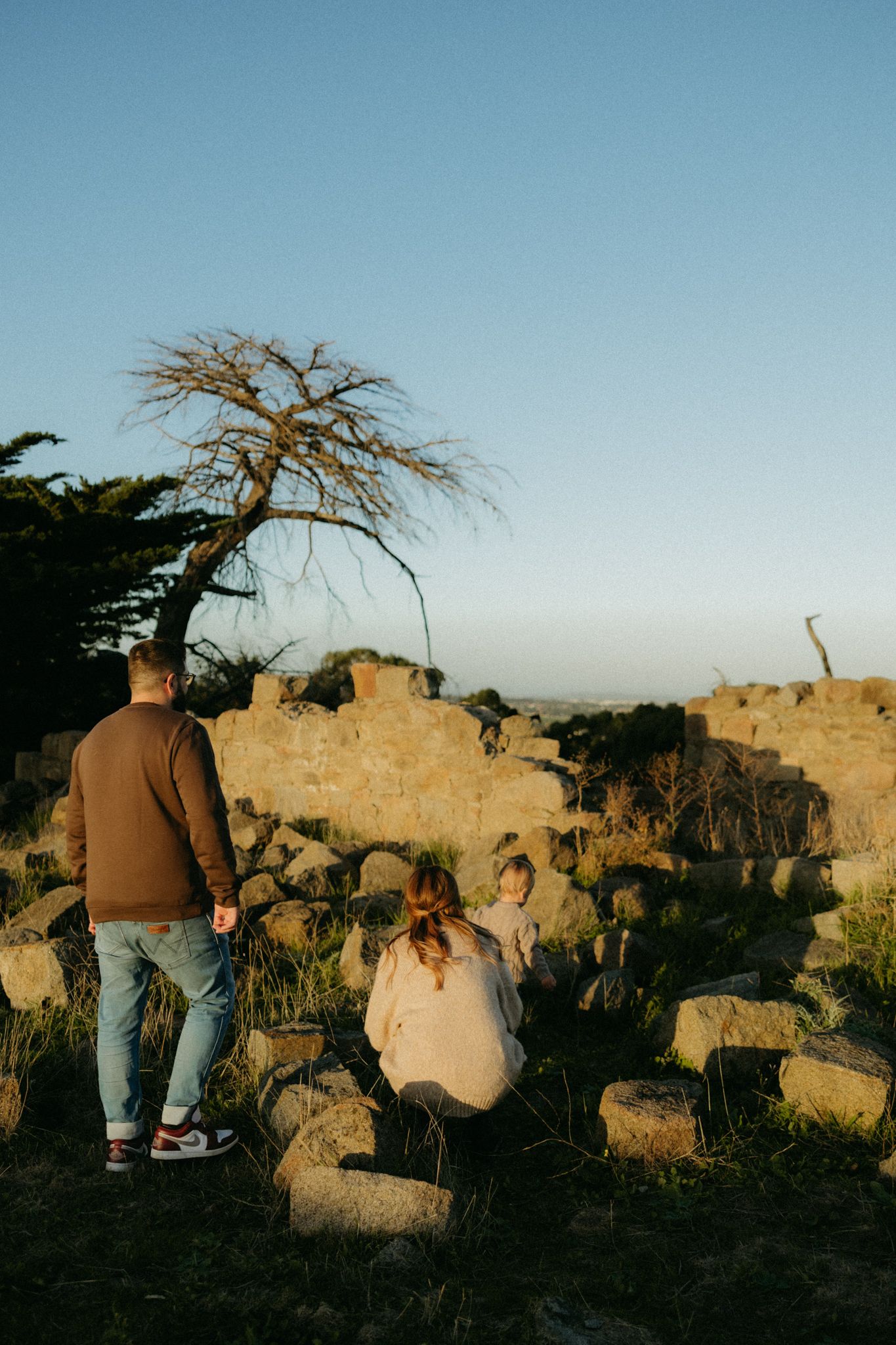 Family of three exploring rocky ruins at golden hour, with a bare tree silhouetted against a clear blue sky.