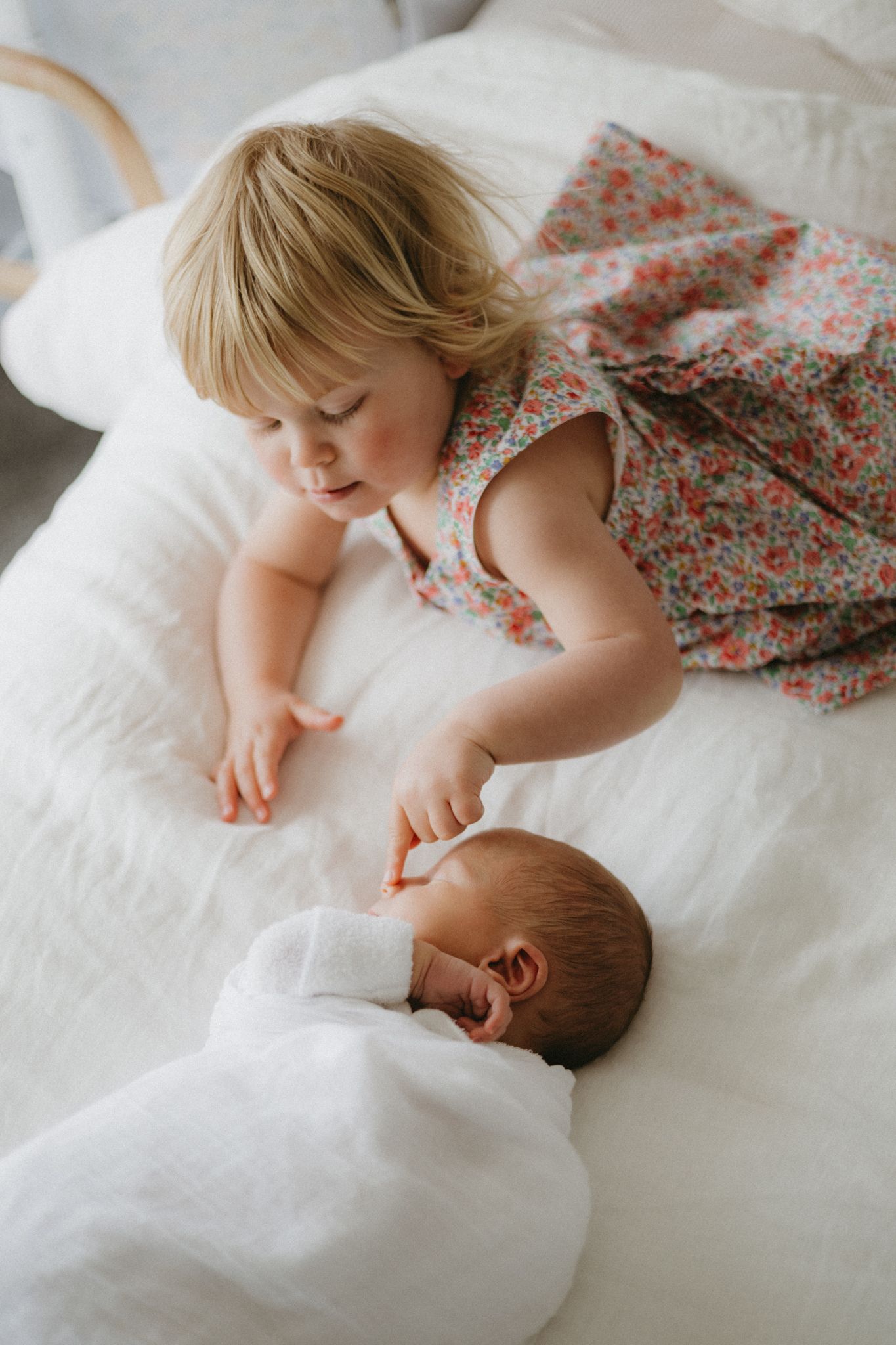 A toddler girl in a floral dress gently touches a swaddled newborn baby lying on a white bed.