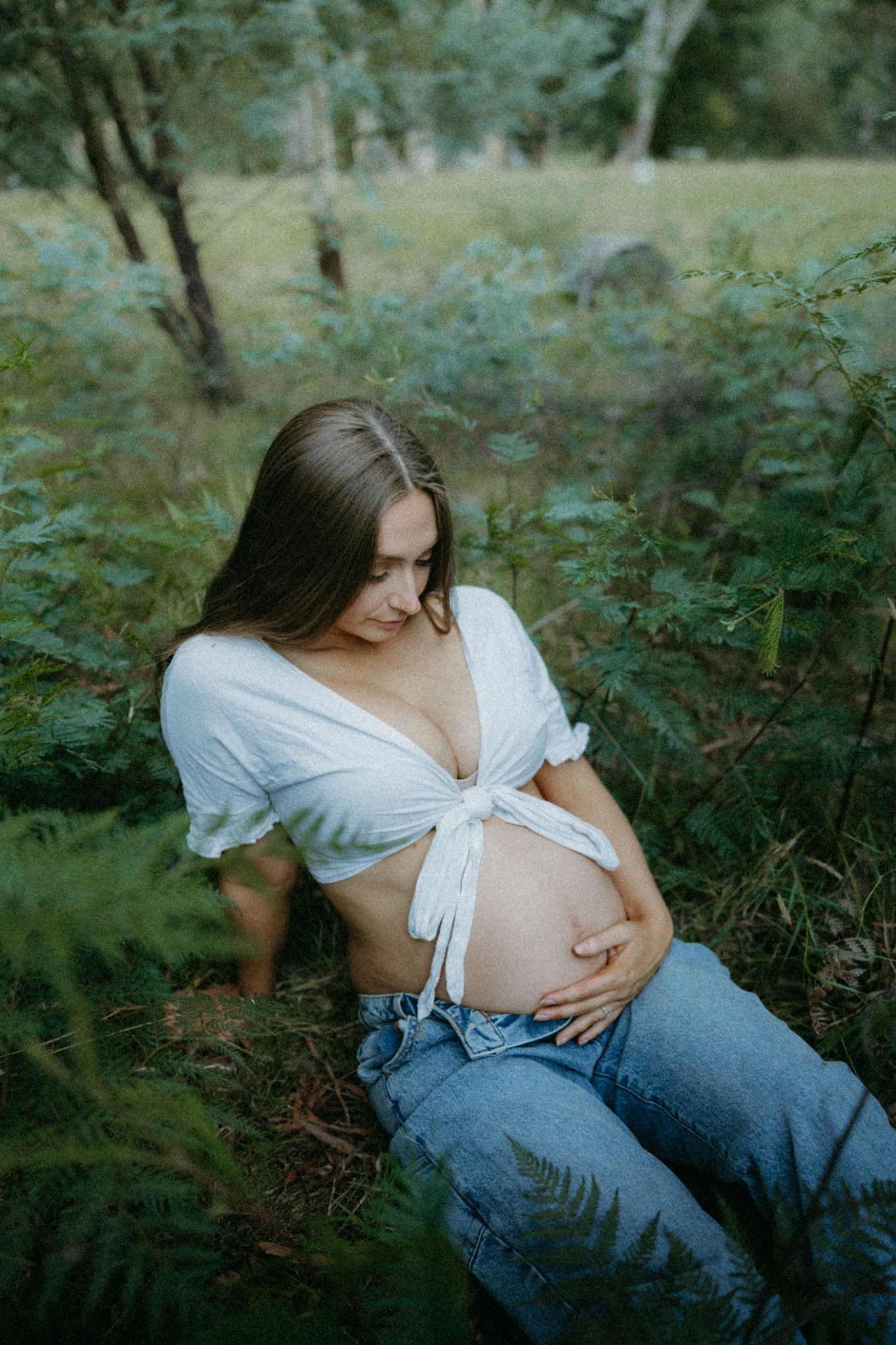 Pregnant woman in white crop top and jeans sitting among ferns, cradling her baby bump in a lush green forest.
