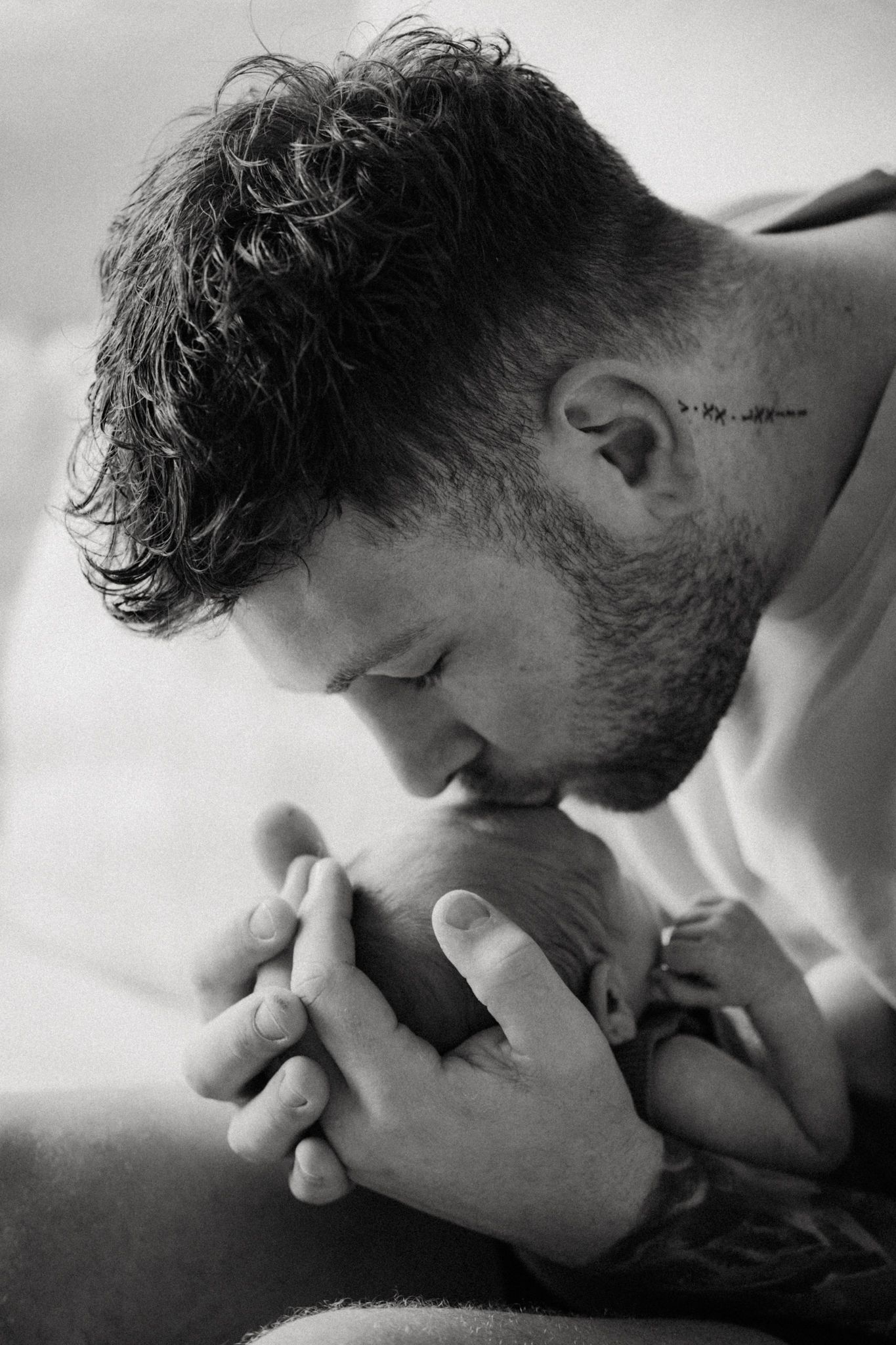 Black and white photo of a bearded man with tattoos tenderly kissing a newborn baby's head while cradling it in his hands.