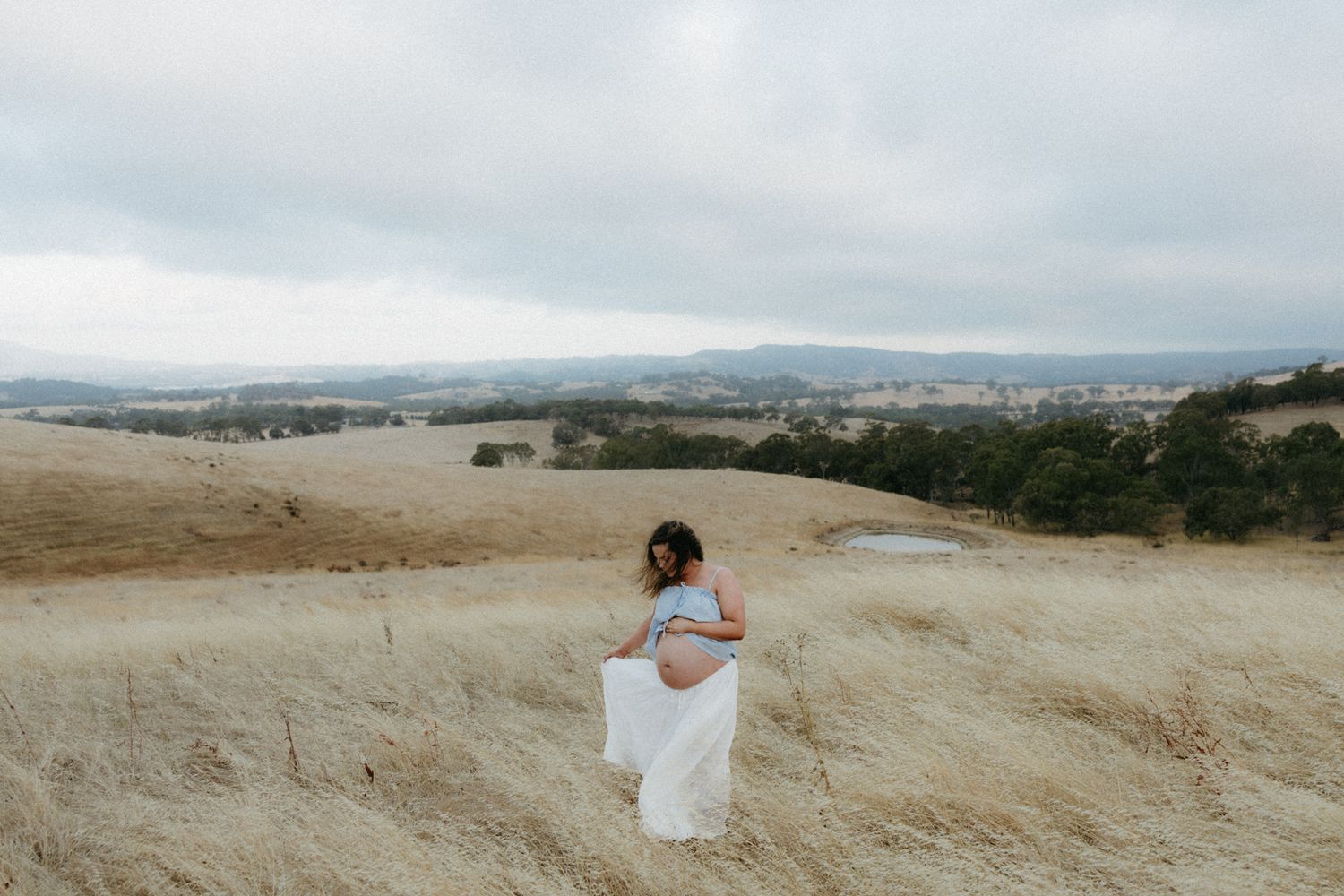 Pregnant woman in white skirt stands in golden field with panoramic valley view under overcast sky.