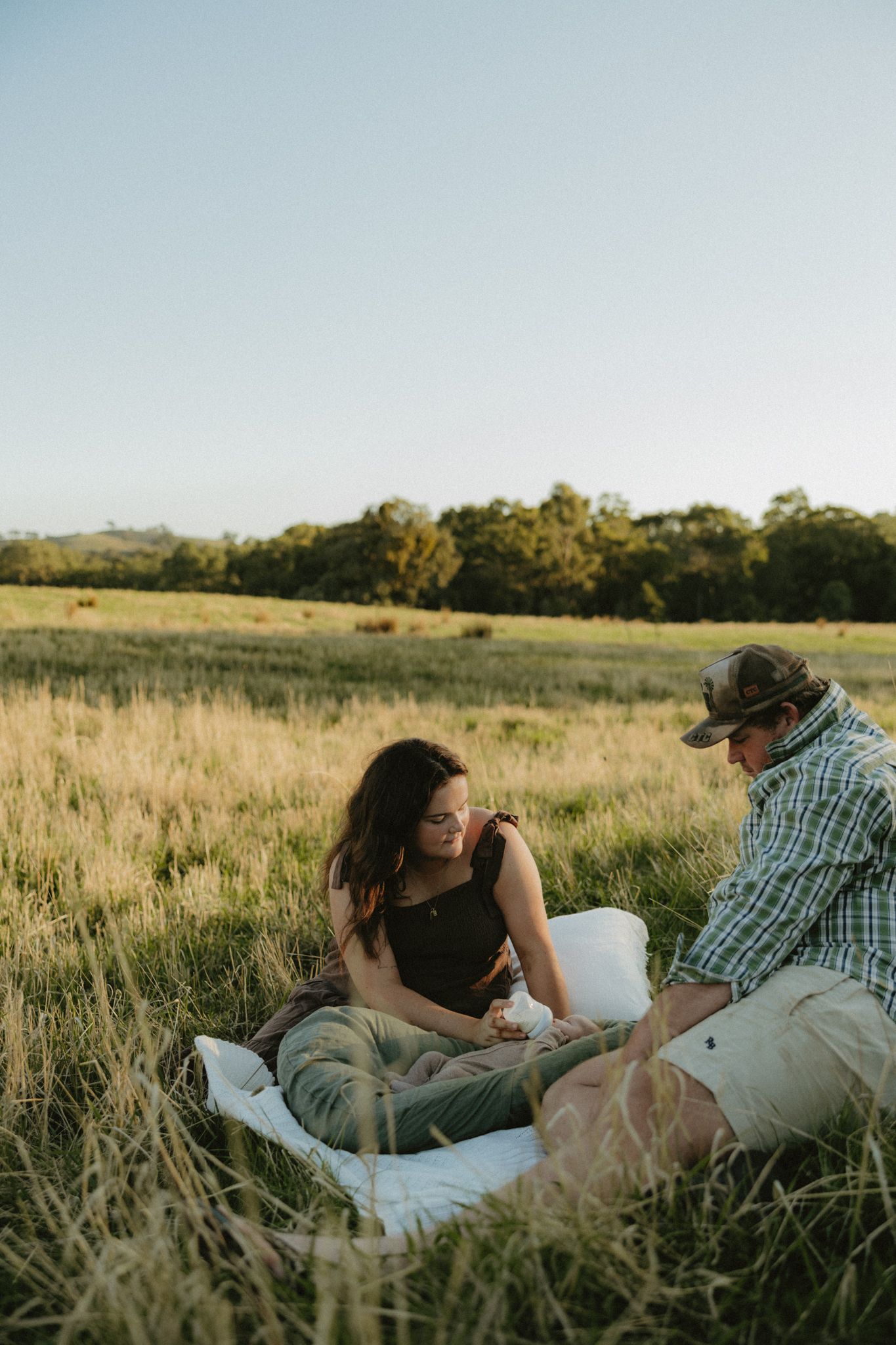 Couple sitting on a blanket in a golden grass field during sunset, sharing an intimate moment in a rural countryside setting.