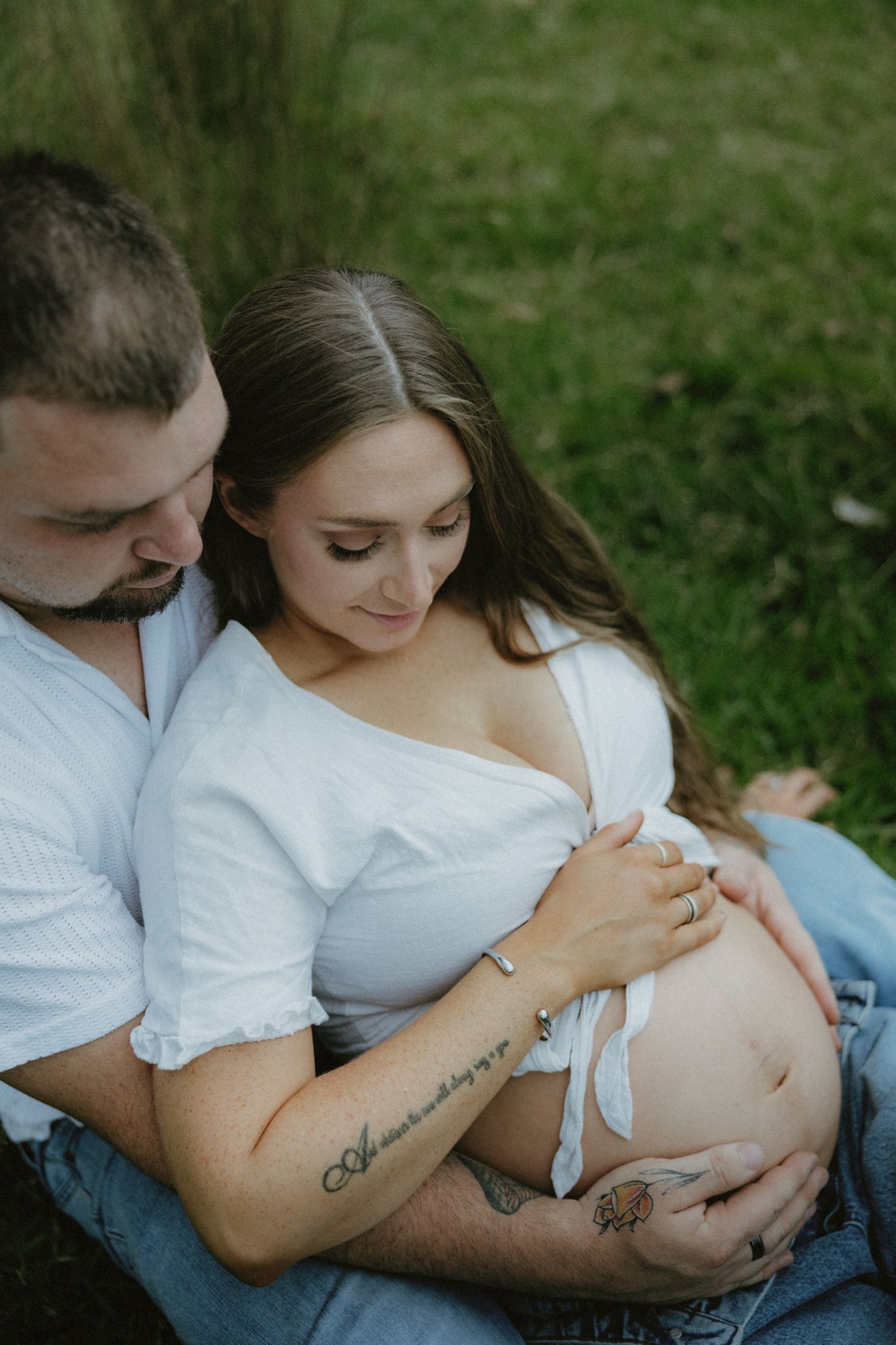 Couple sitting outdoors in grass, pregnant woman in white top cradling baby bump with partner embracing her tenderly.