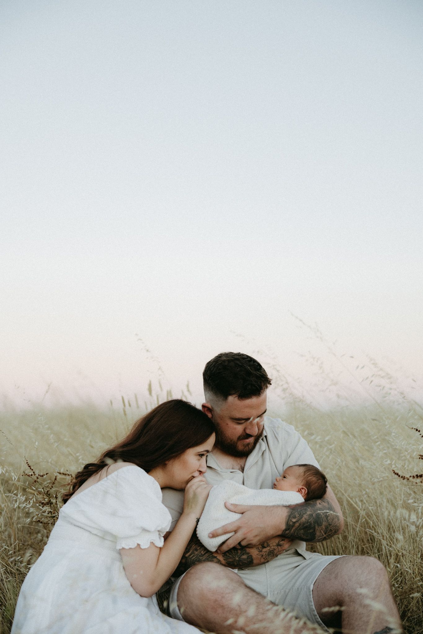 A couple sits in a golden field at dusk, tenderly gazing at their newborn baby wrapped in white.