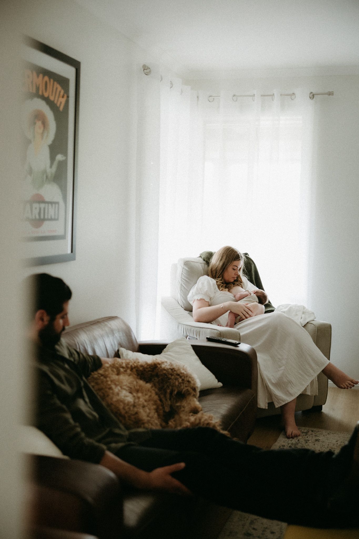 A woman in white dress nursing newborn in recliner while man relaxes on leather sofa with fluffy dog nearby.