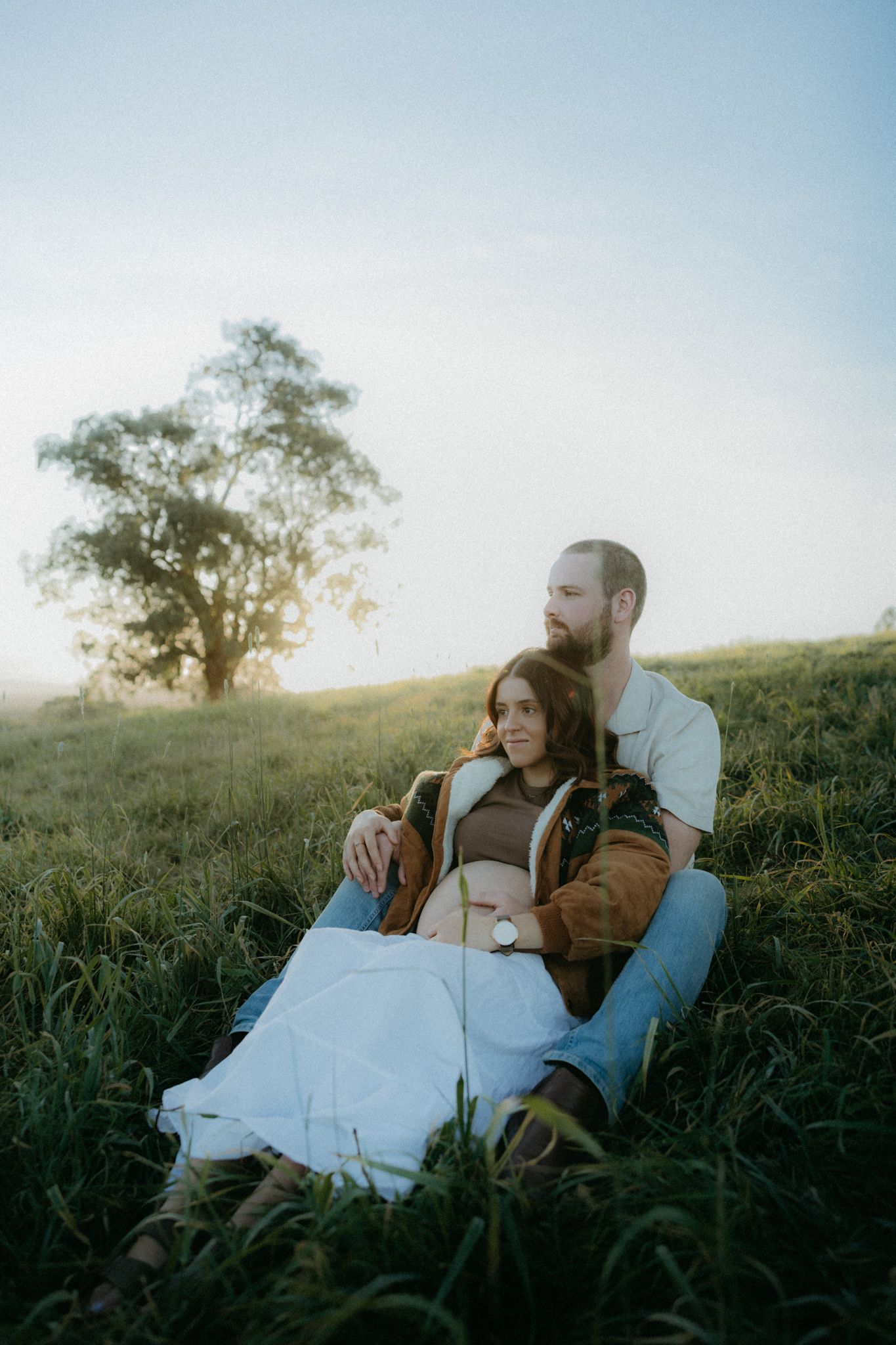 A couple sits together on a grassy hillside at dusk, with a lone tree silhouetted against a misty sky.