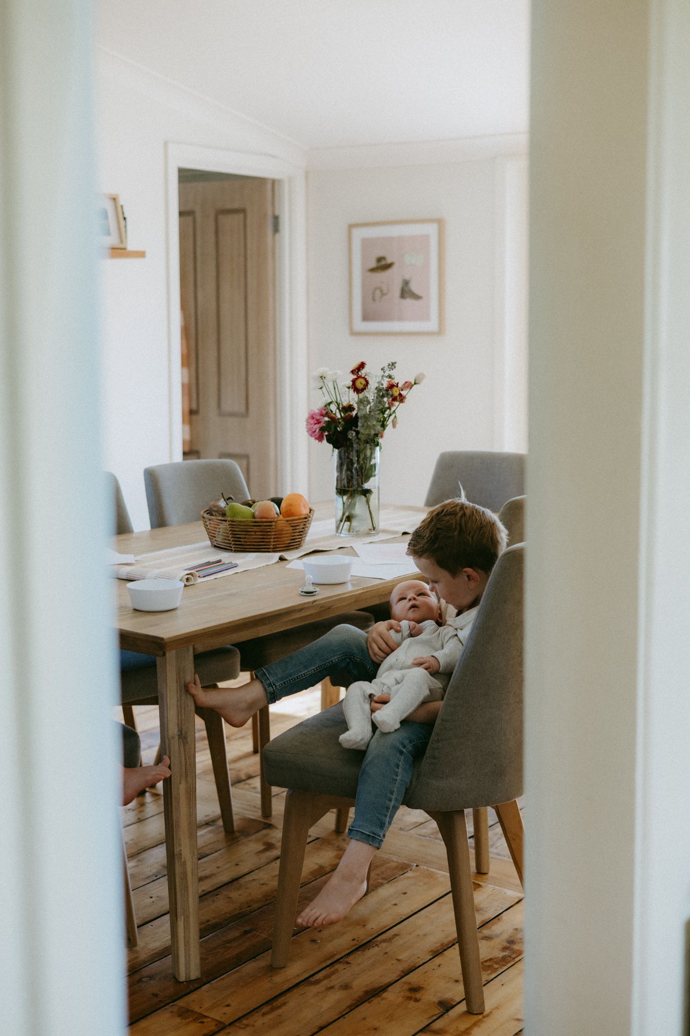 Child sitting in dining chair holding a newborn baby in a bright, modern home with wooden floors and fresh flowers.
