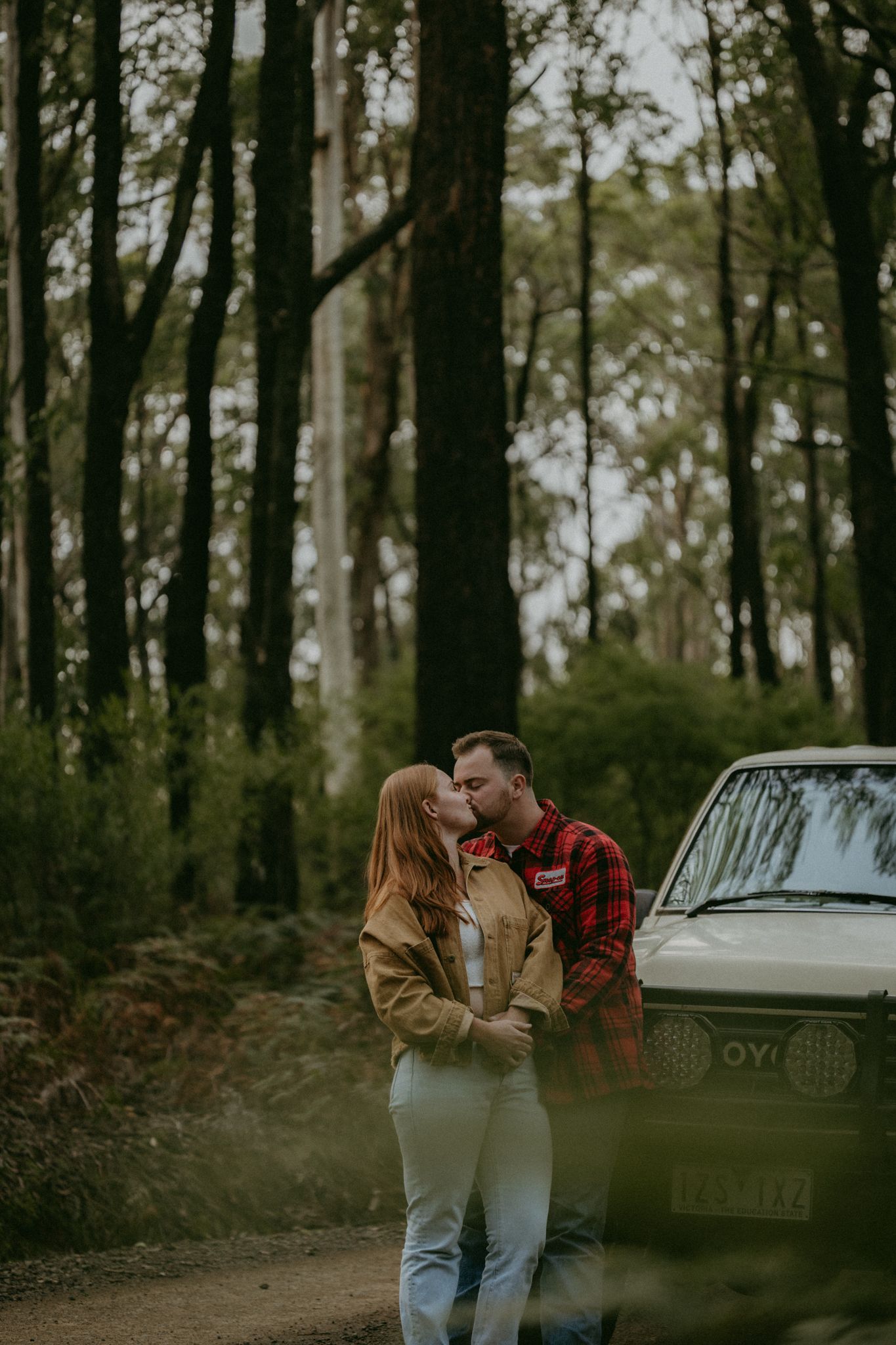 Couple sharing a romantic kiss in a forest beside a vintage car, wearing casual jackets.
