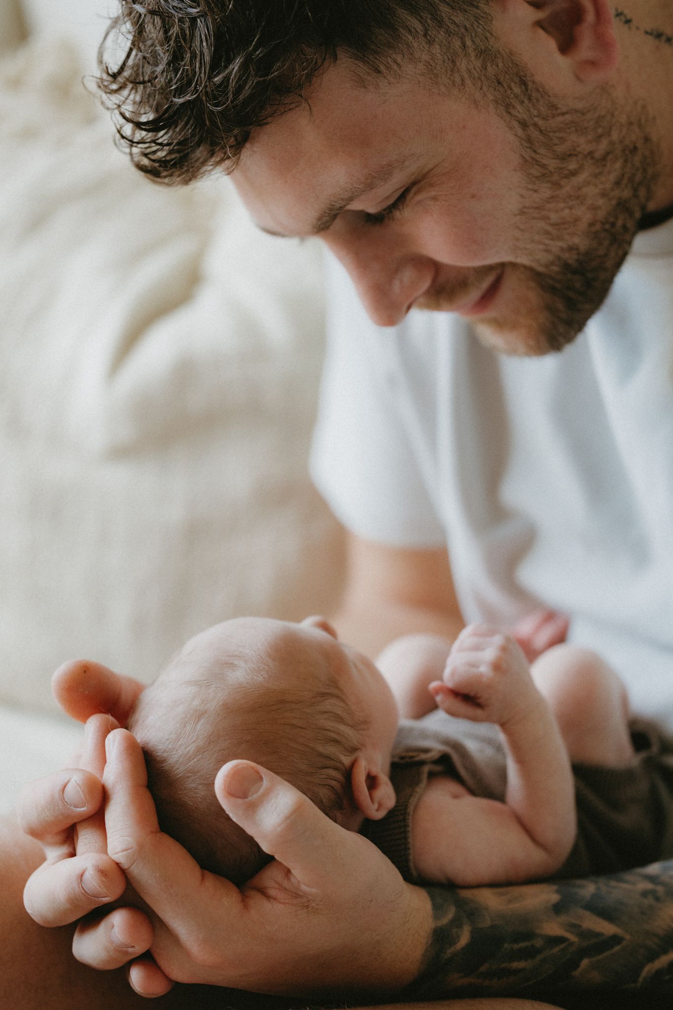 A tattooed father tenderly cradling his newborn baby in his hands, smiling down with love.