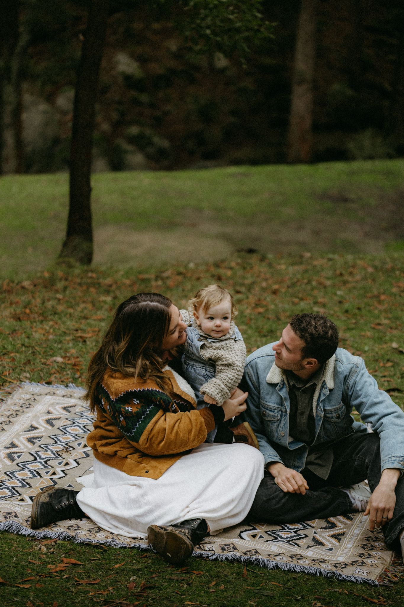 Family of three sitting on a patterned blanket in a lush green park, parents smiling at their toddler.