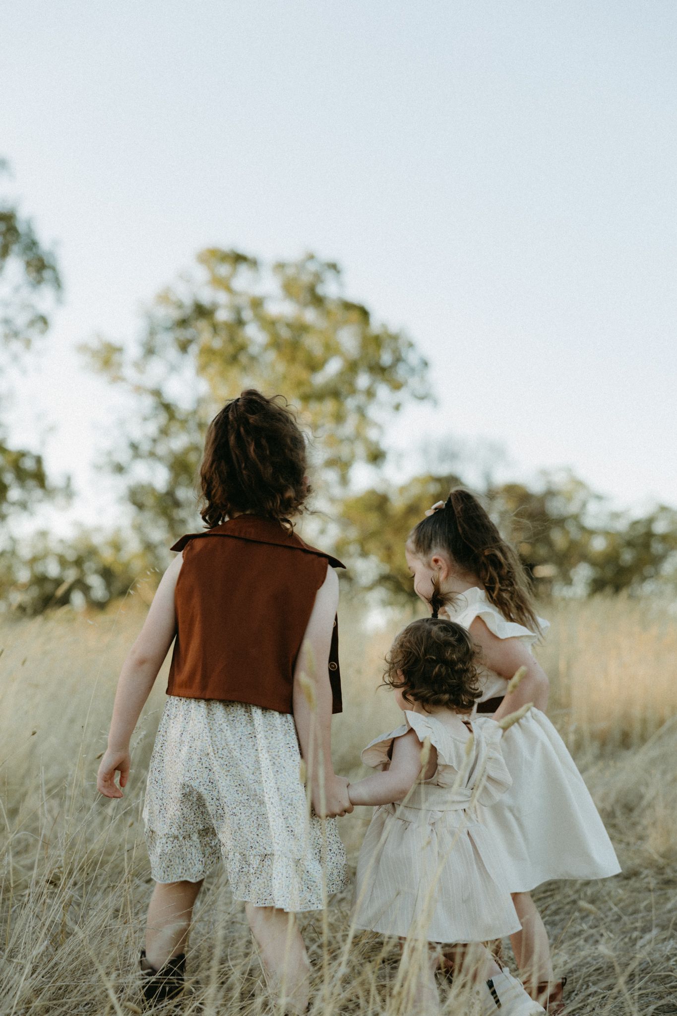 Three young girls in white and rust dresses walk hand-in-hand through golden grass fields with trees in background.