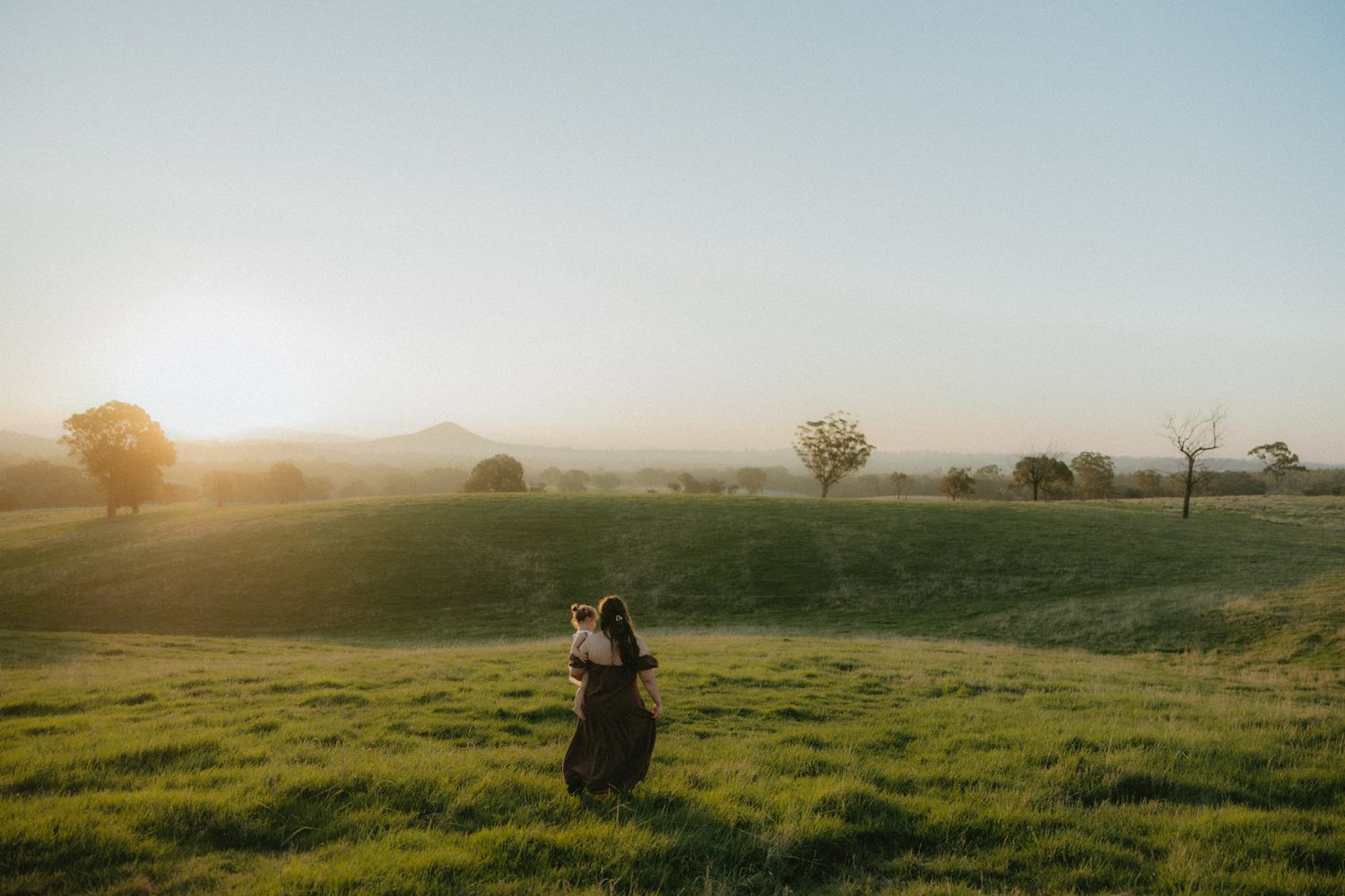 Person walking through lush green fields at golden hour with misty mountains and trees in the background.