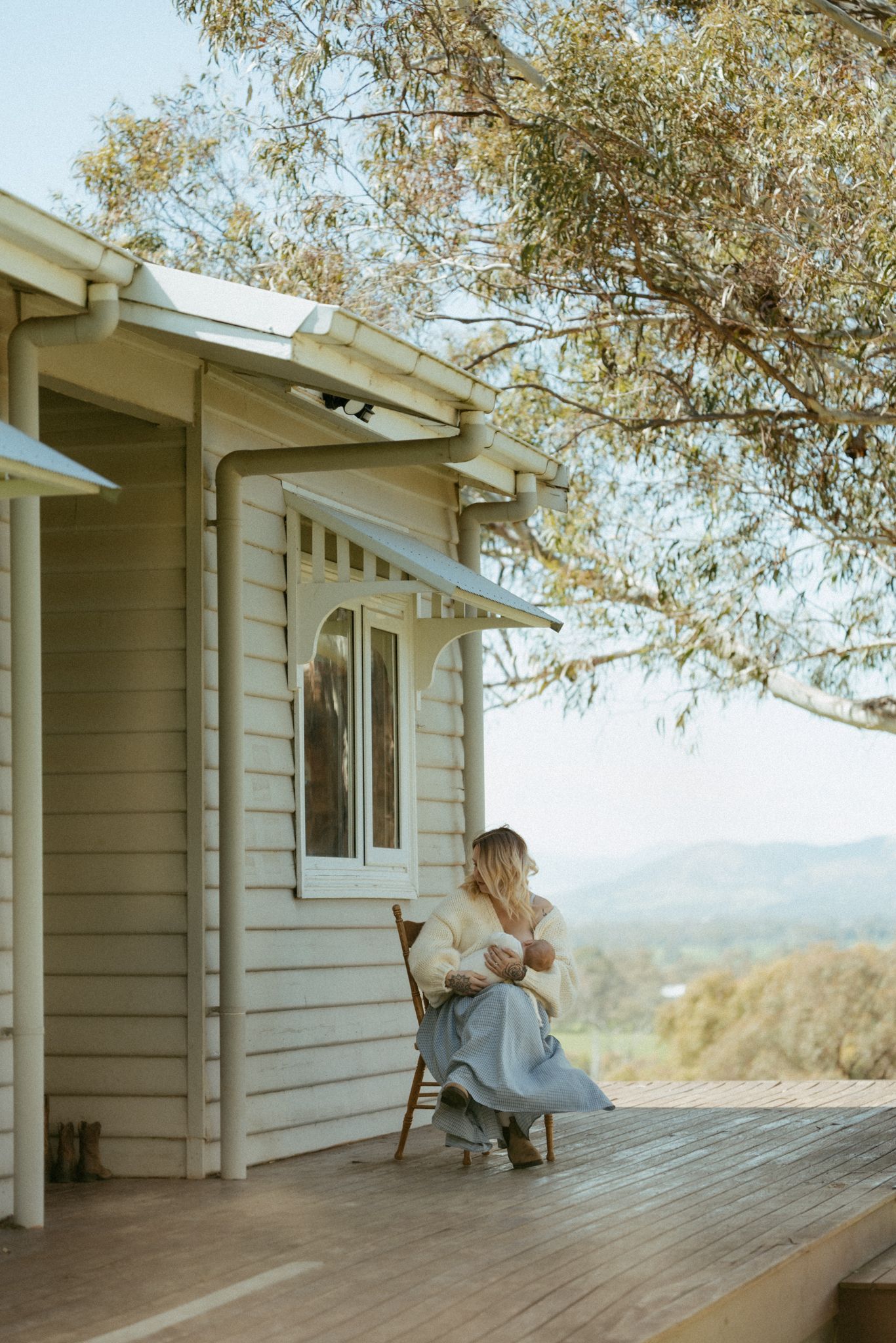 Woman sitting on a wooden deck of a white cottage, nursing a baby with scenic hills and eucalyptus trees in background.