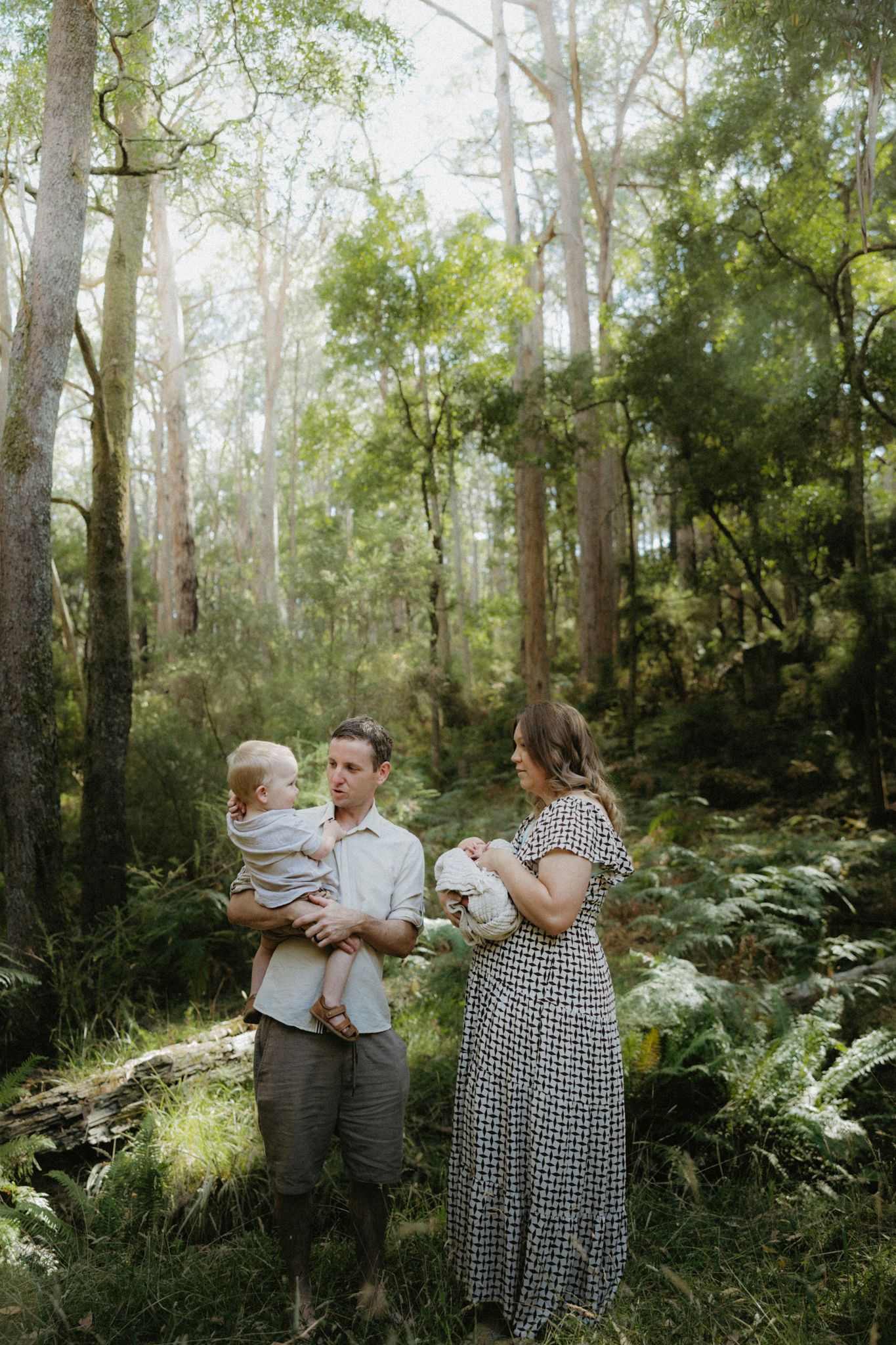 A family of three stands in a lush green forest, father holding toddler while mother cradles newborn baby.