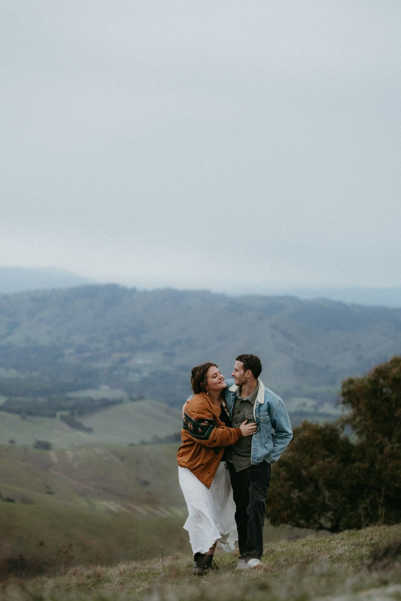 Couple embraces on misty hilltop, woman in rust jacket and white skirt leaning into partner with scenic valley views behind.