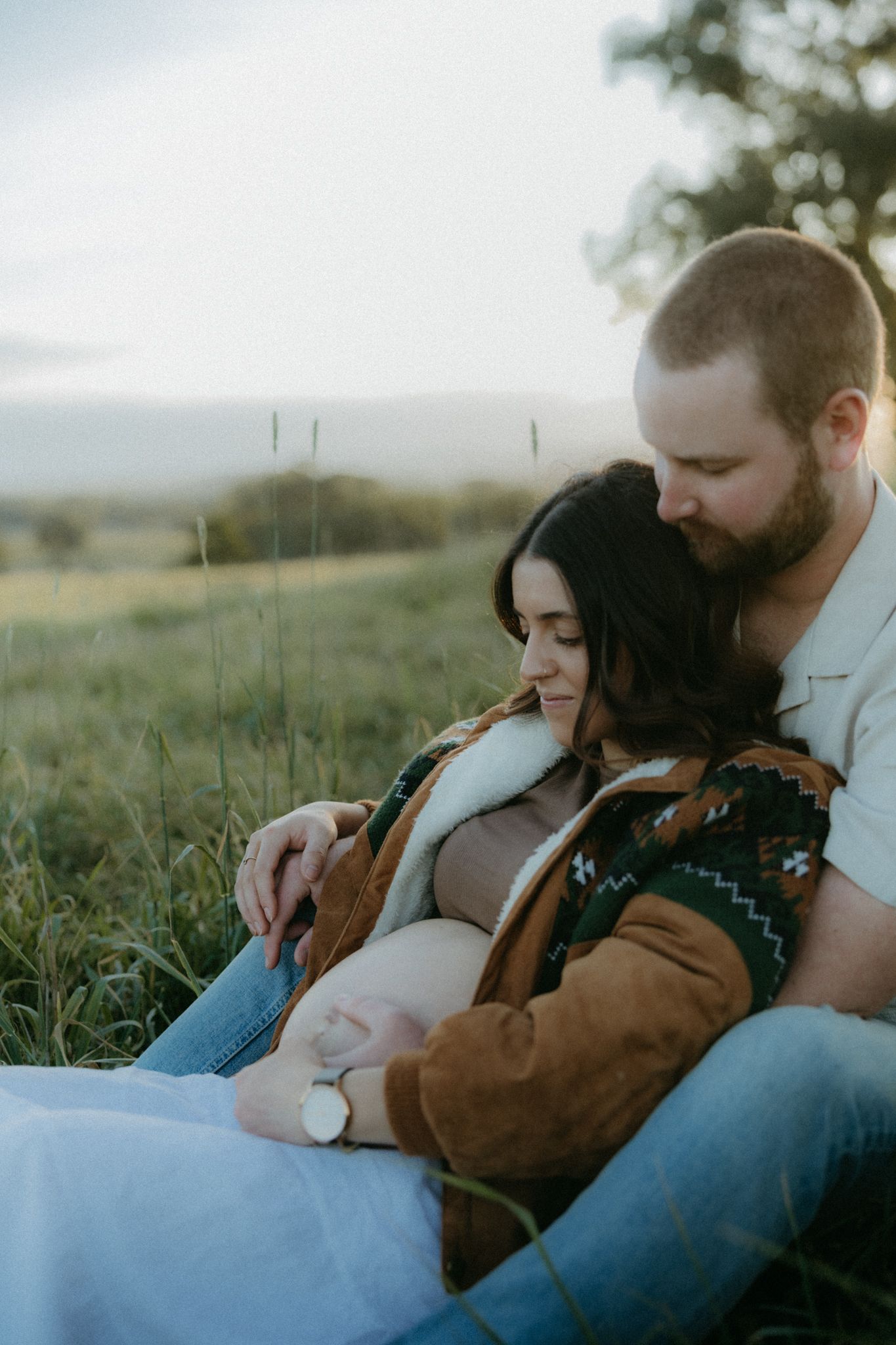 Couple sitting in a field, man embracing pregnant woman from behind, both looking down at her baby bump.