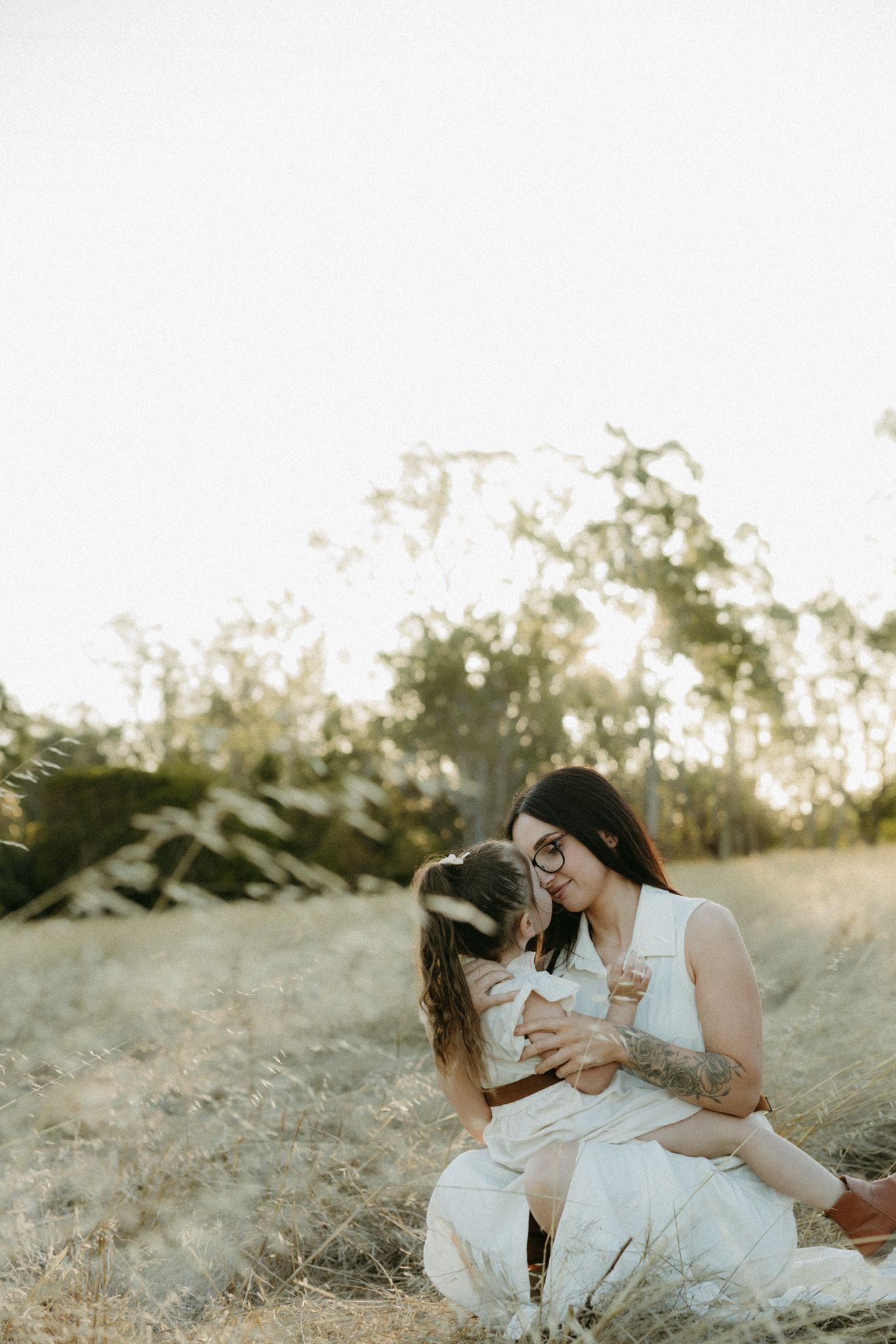 Mother and young daughter share a tender kiss while sitting in a golden field at sunset, both wearing white.