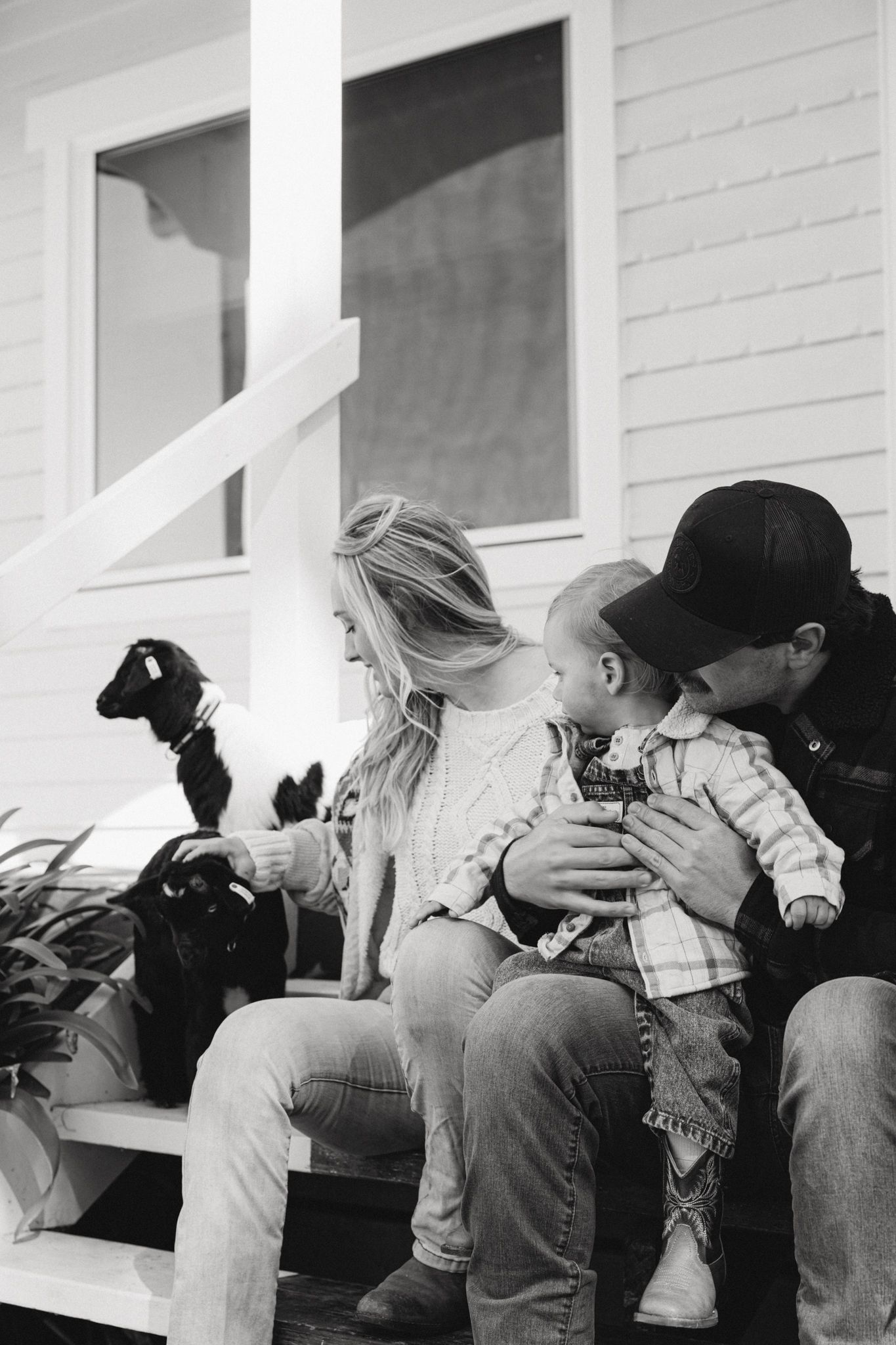 Black and white photo of a family sitting on porch steps, parents with baby and small black dog.