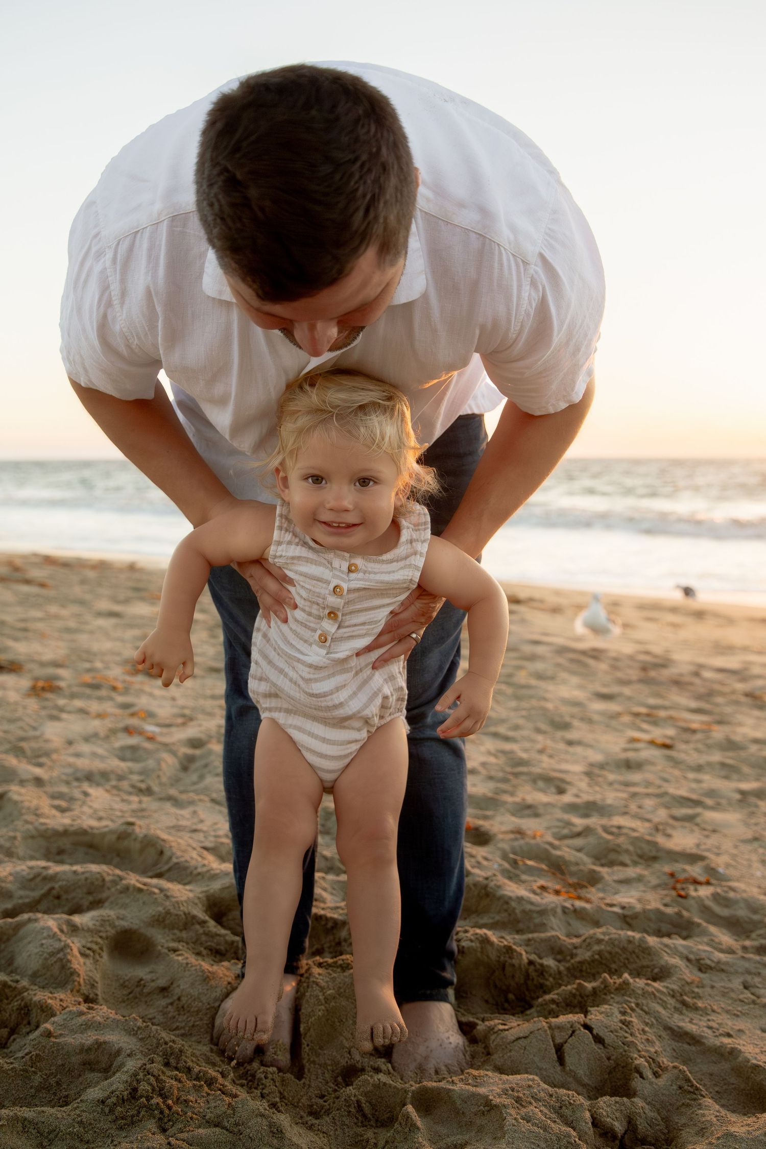 Parent helping small child stand on sandy beach during sunset.