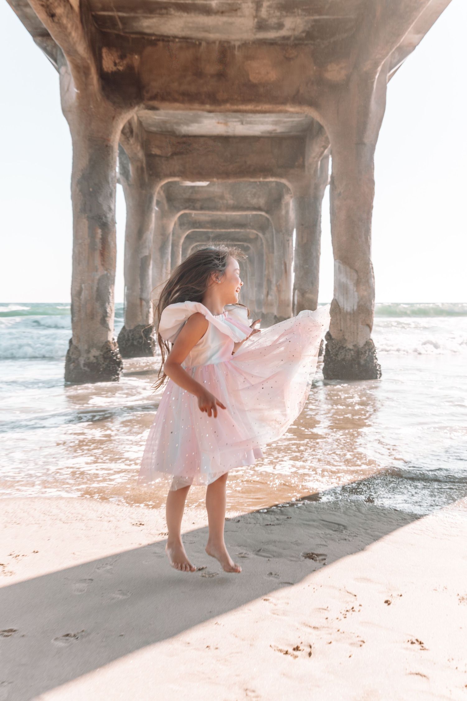 Person in pink dress twirling under a pier at the beach with sunlight streaming through.