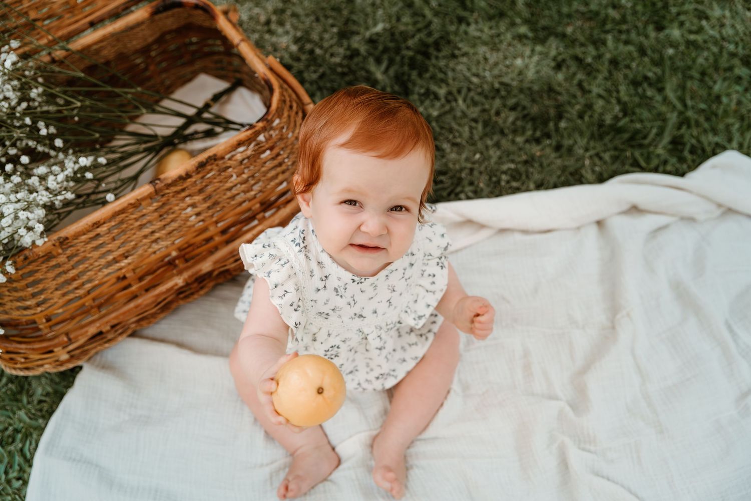 Baby in floral dress reaches for oranges while lying on white blanket outdoors.